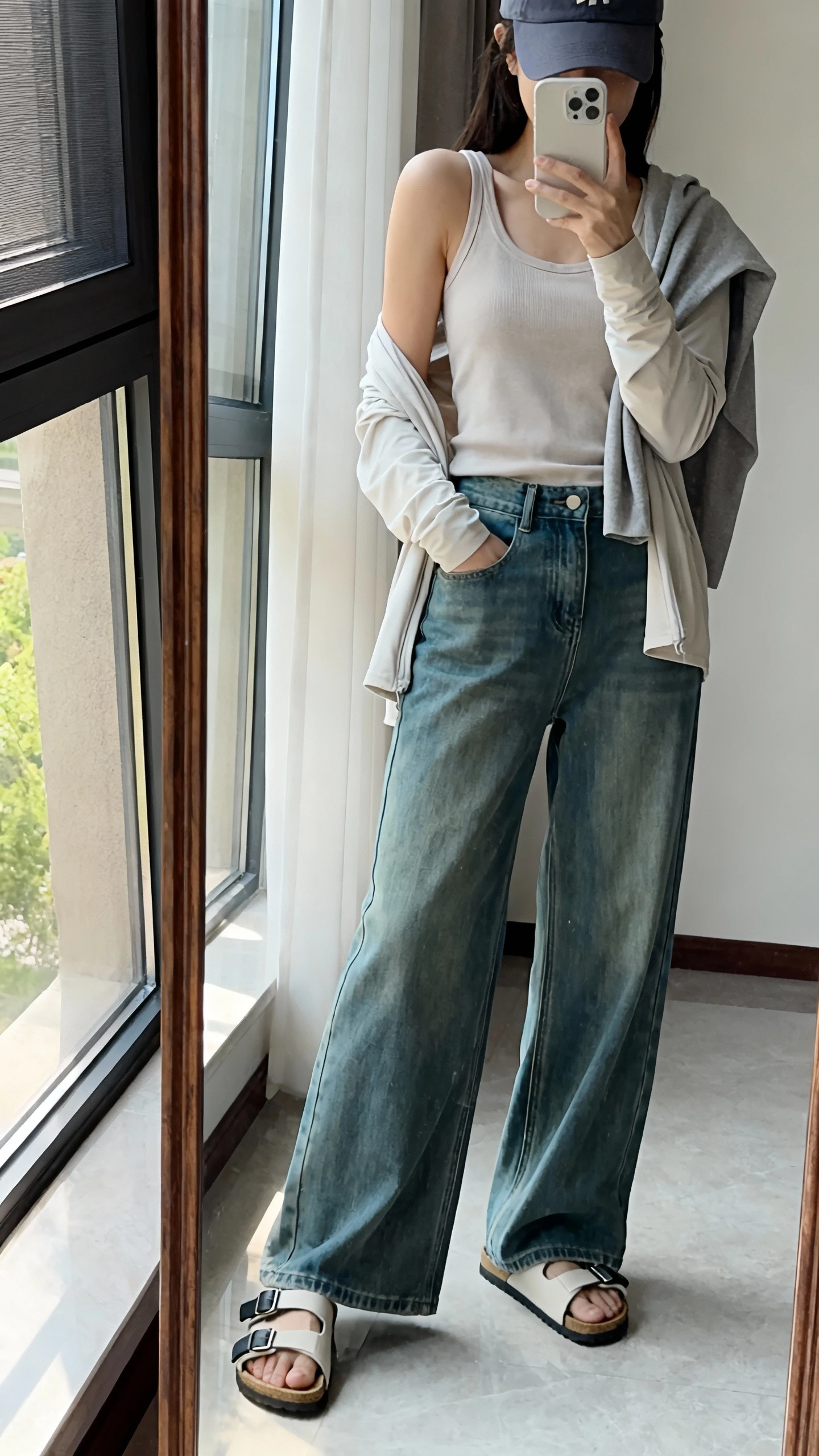 iPhone side-angle mirror selfie of a woman wearing wide-leg jeans, sporty Velcro-strap sandals, a relaxed tank, lightweight cardigan draped over shoulders, and a baseball cap, face not visible, full-length mirror near a window with soft morning light, iPhone photo quality.