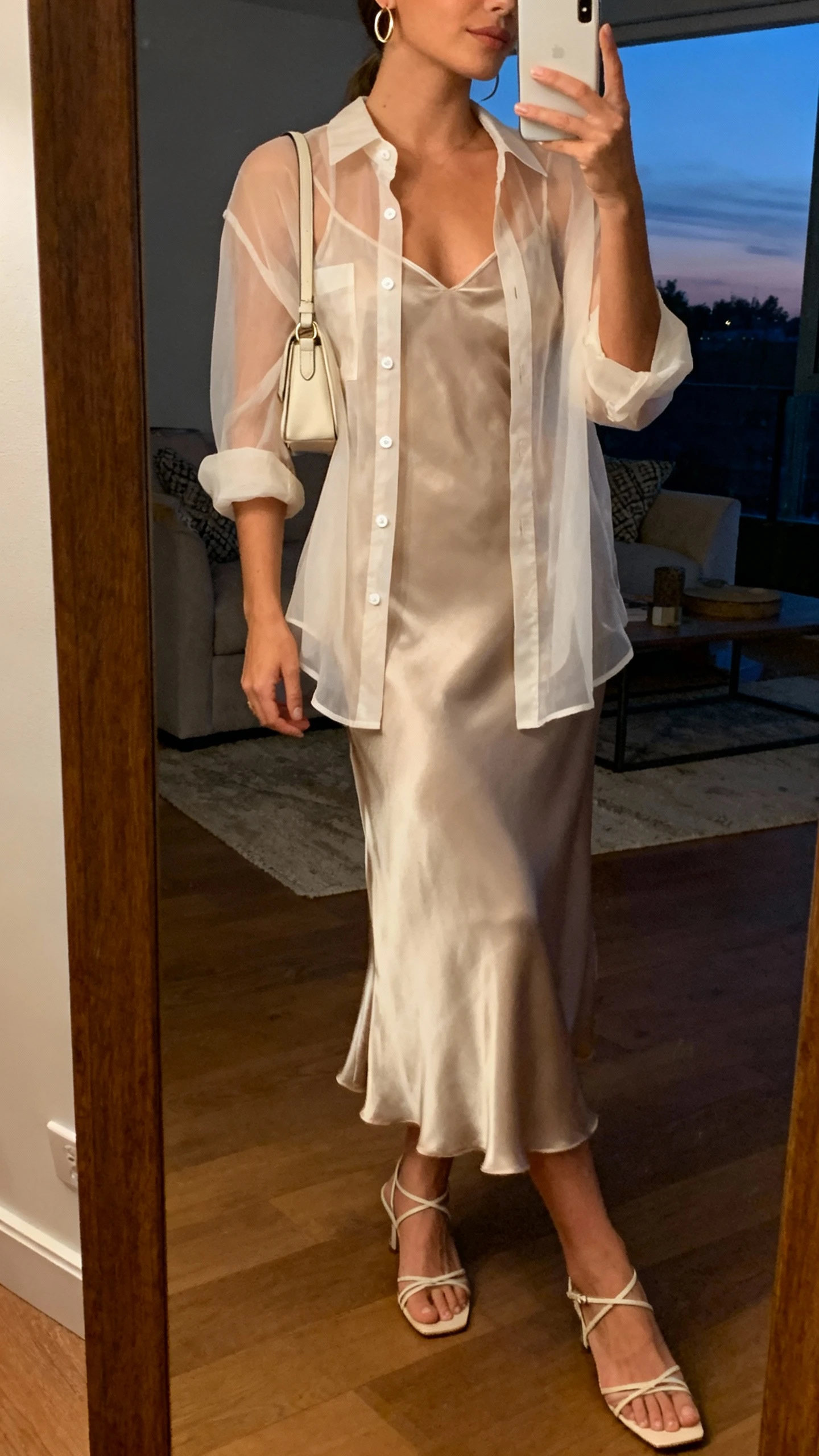 iPhone side-angle mirror selfie of a woman wearing a silky slip dress layered with a sheer button-up shirt, delicate strappy heels, small shoulder bag, thin hoop earrings, face not visible, living room full-length mirror, warm evening indoor lighting, iPhone photo quality.