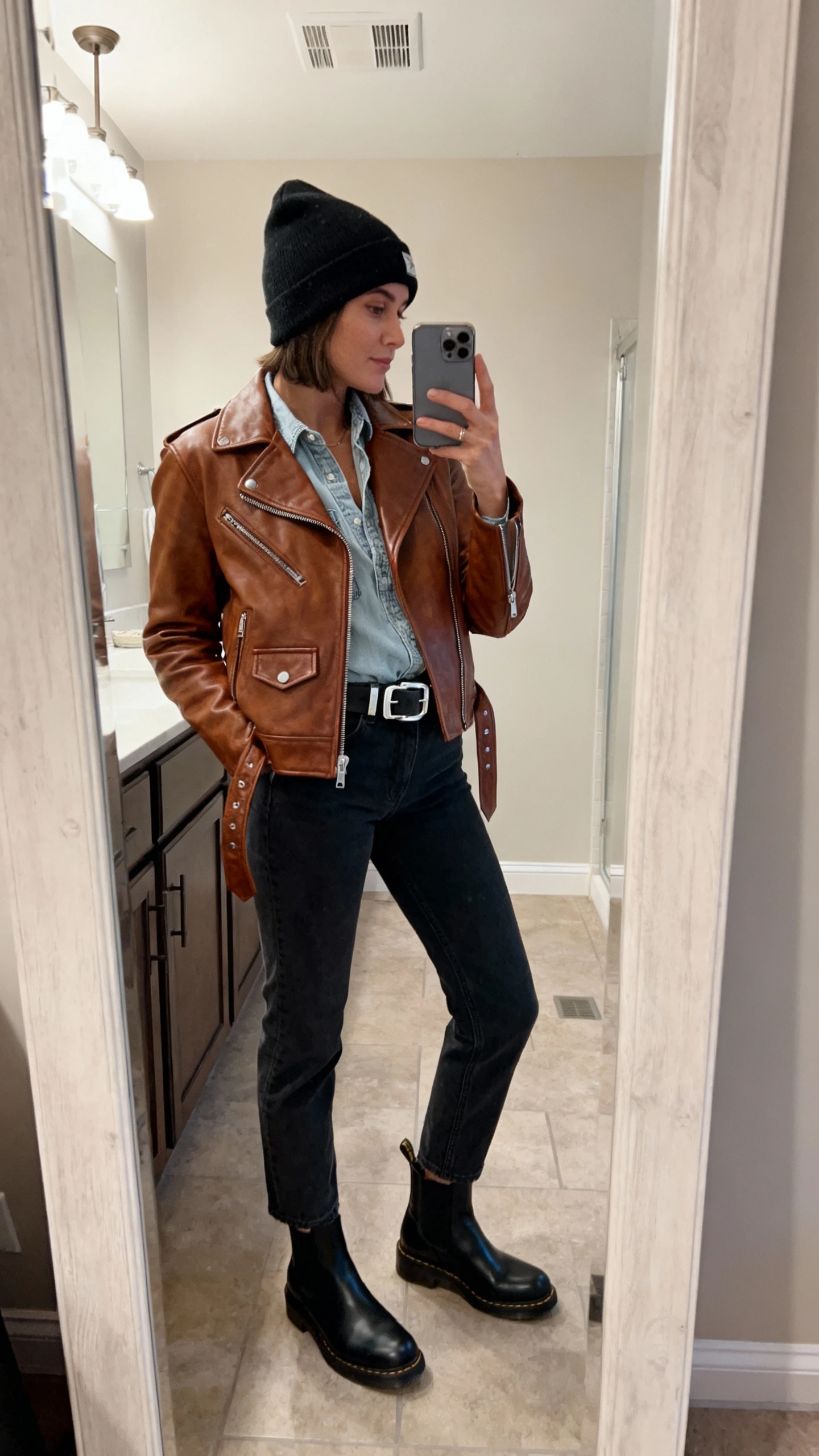 iPhone side-angle mirror selfie of a woman wearing a brown leather biker jacket with a light denim shirt, dark straight-leg jeans, black Chelsea boots, a black beanie, and a silver-buckle belt, face not visible, bathroom mirror, overhead indoor lighting, iPhone photo quality.