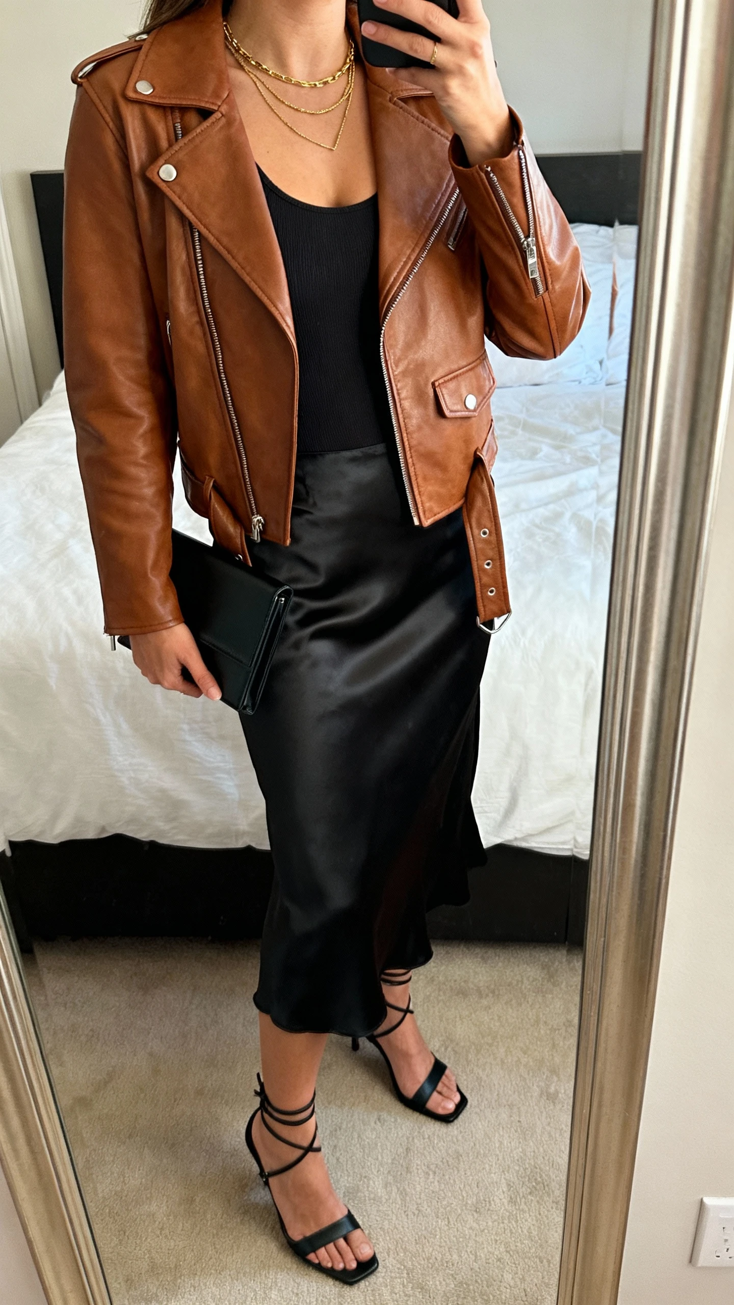 iPhone side-angle mirror selfie of a woman wearing a brown leather moto jacket over a black fitted bodysuit, black satin midi slip skirt, strappy black heels, a slim black clutch, and delicate gold layered necklaces; face not visible, cropped at shoulders, bedroom mirror, warm indoor lighting, iPhone photo quality.