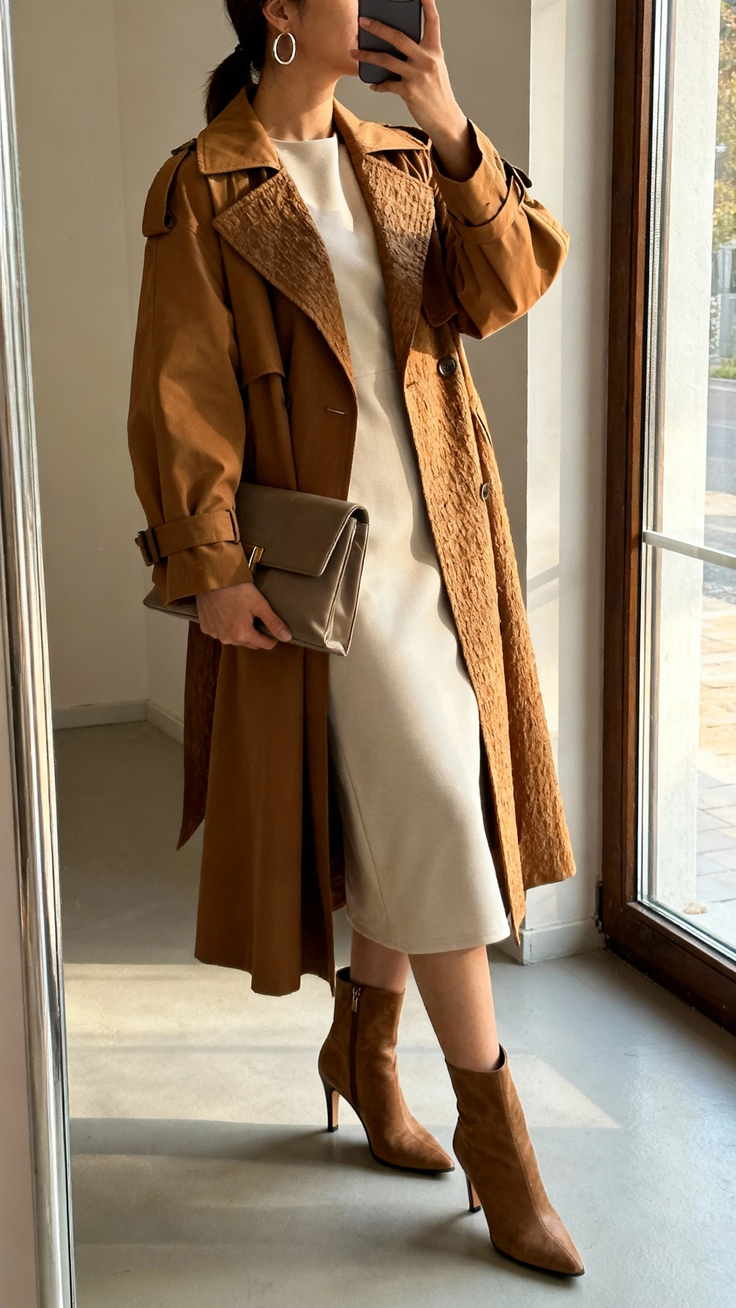 iPhone side-angle mirror selfie of a woman wearing a minimal midi dress under statement outerwear (bold trench or textured coat), heeled ankle boots, structured clutch, simple hoop earrings, face not visible, full-length mirror near window, soft afternoon light, iPhone photo quality