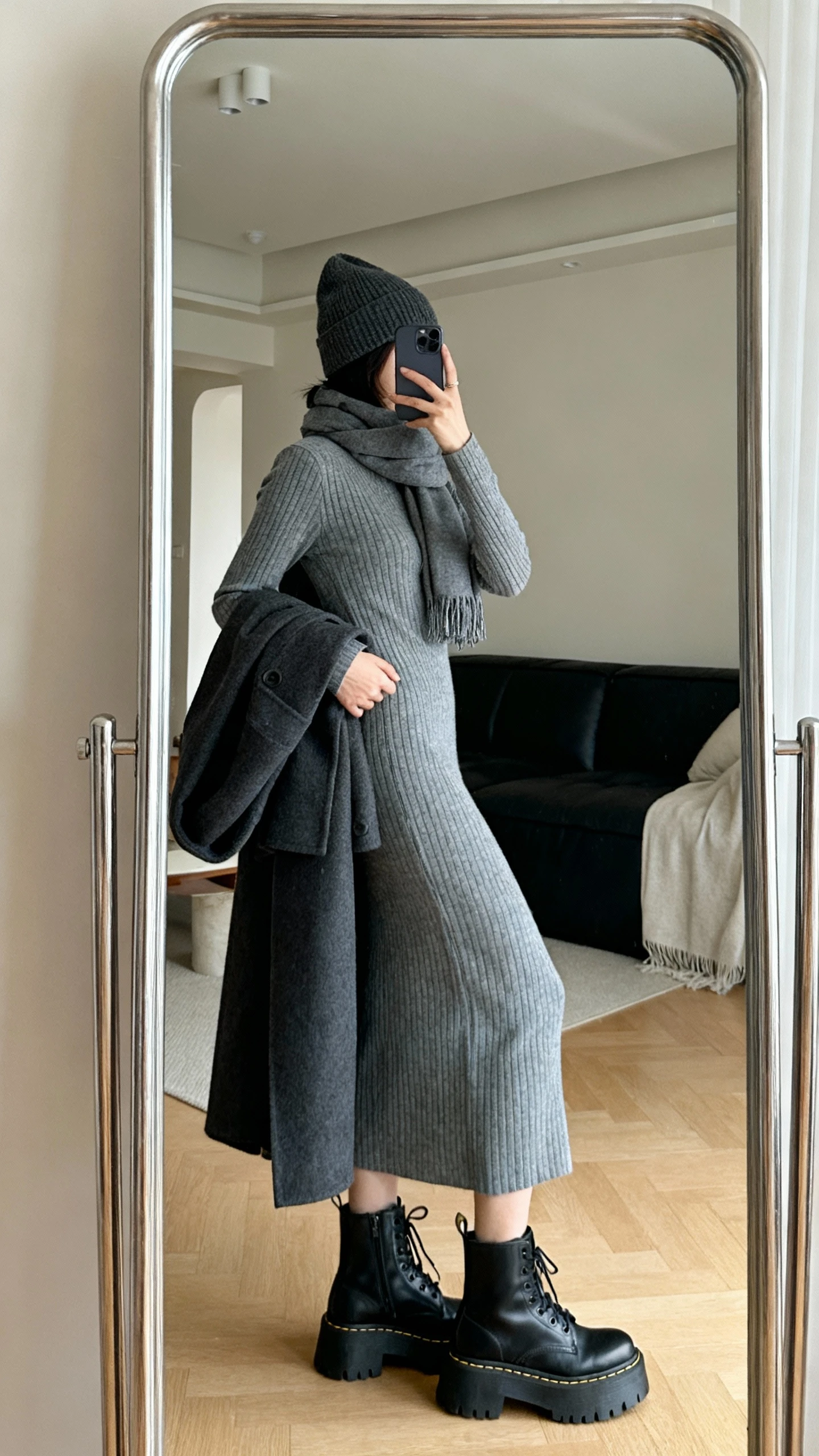 iPhone side-angle mirror selfie of a woman wearing a ribbed knit midi dress in heather gray, chunky black commuter lace-up boots, long coat draped over arm, beanie, and a scarf, face not visible, living room full-length mirror, soft indoor lighting, iPhone photo quality.