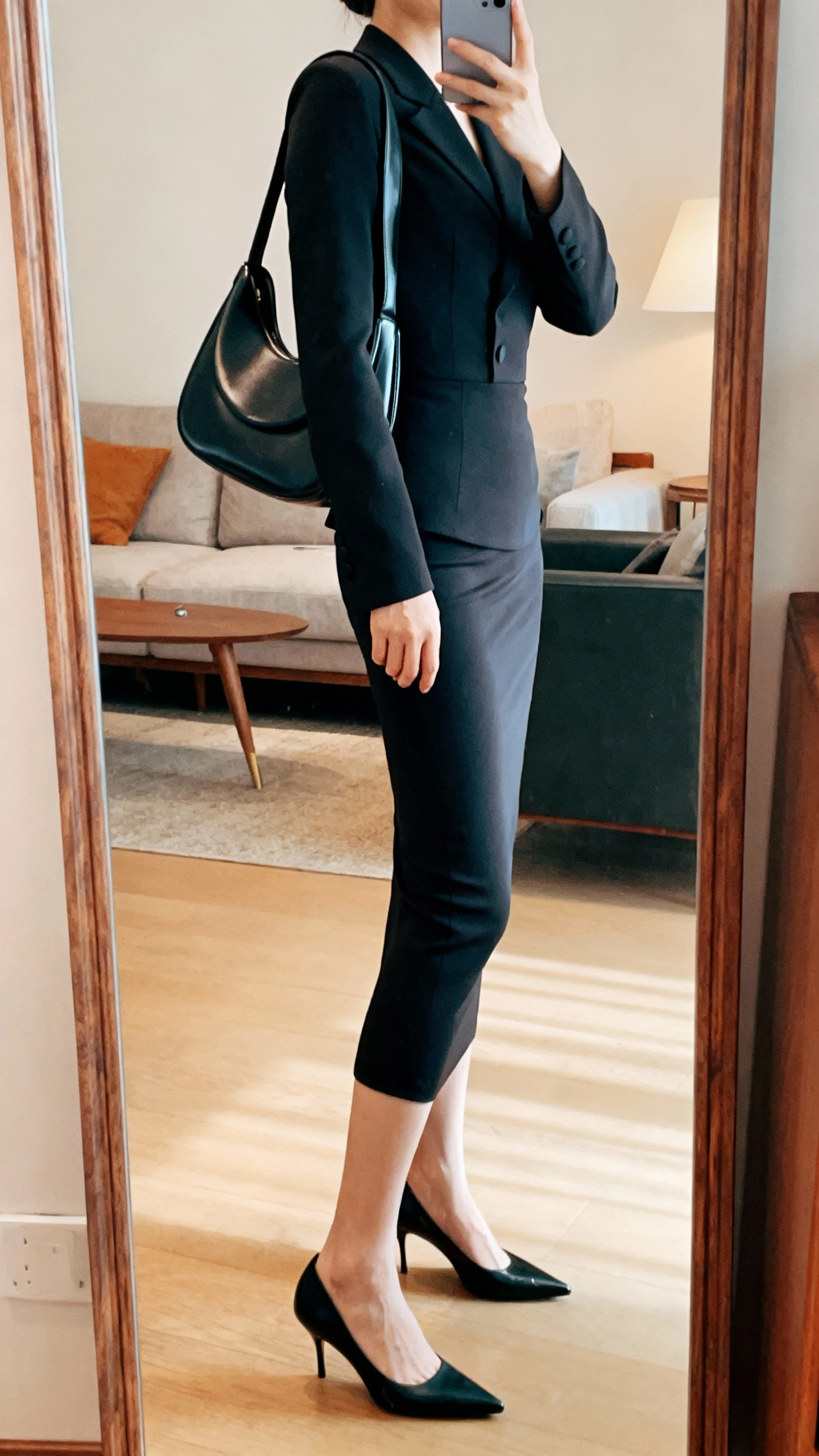 iPhone side-angle mirror selfie of a woman wearing a fitted after-dark monochrome set: tailored fitted top, slim skirt, pointed heels, and a sculptural shoulder bag; face not visible, living room full-length mirror, warm indoor lighting, iPhone photo quality.