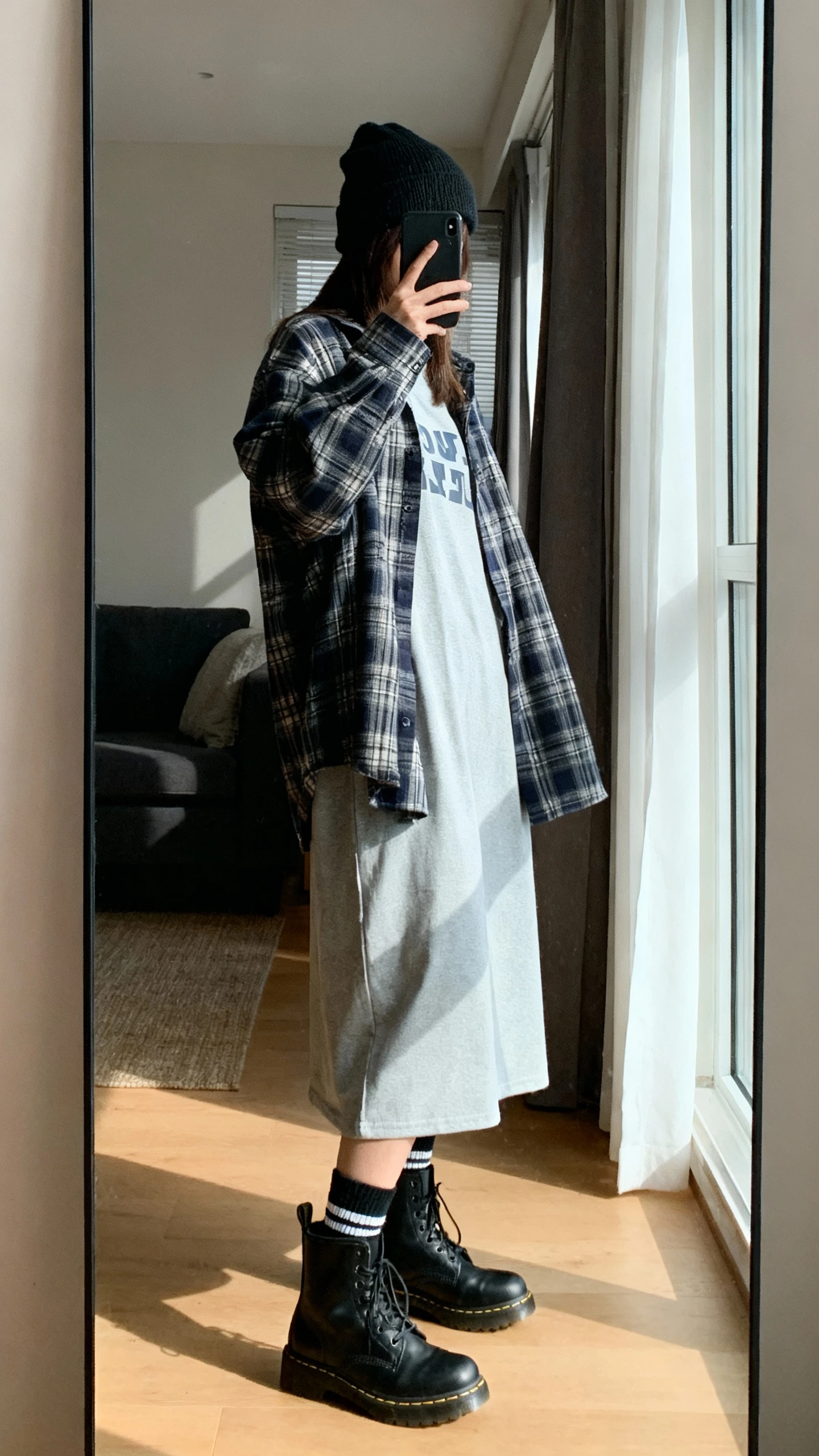 iPhone side-angle mirror selfie of a woman wearing a long oversized T-shirt layered under an open plaid flannel, black combat boots, crew socks peeking, and a beanie, face not visible, living room full-length mirror with afternoon window light, iPhone photo quality.