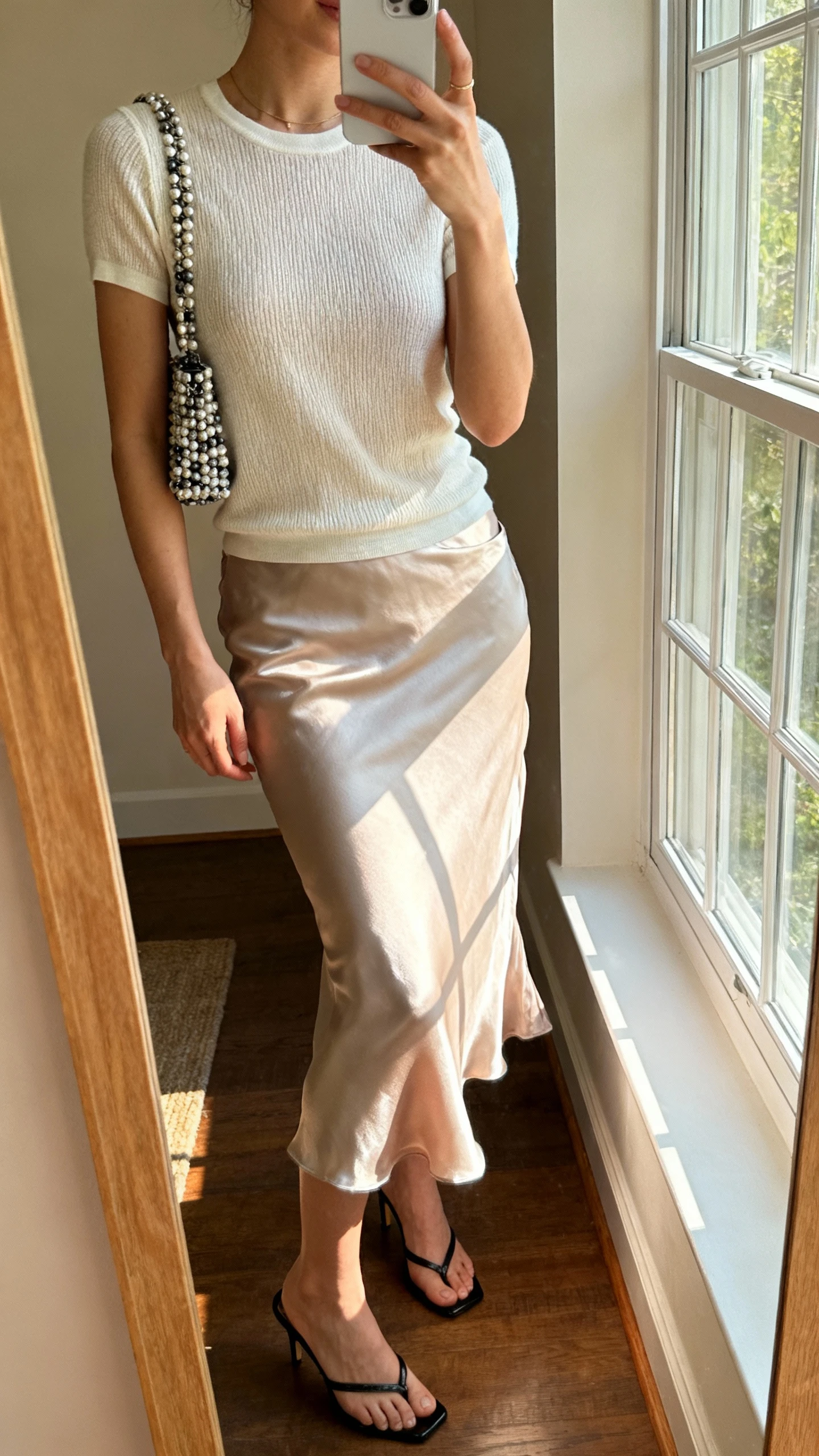 iPhone side-angle mirror selfie of a woman wearing a satin slip skirt with a fitted knit tee, a beaded crossbody bag, and kitten-heel mules, face not visible, cropped at neck, warm indoor lighting by window, iPhone photo quality.