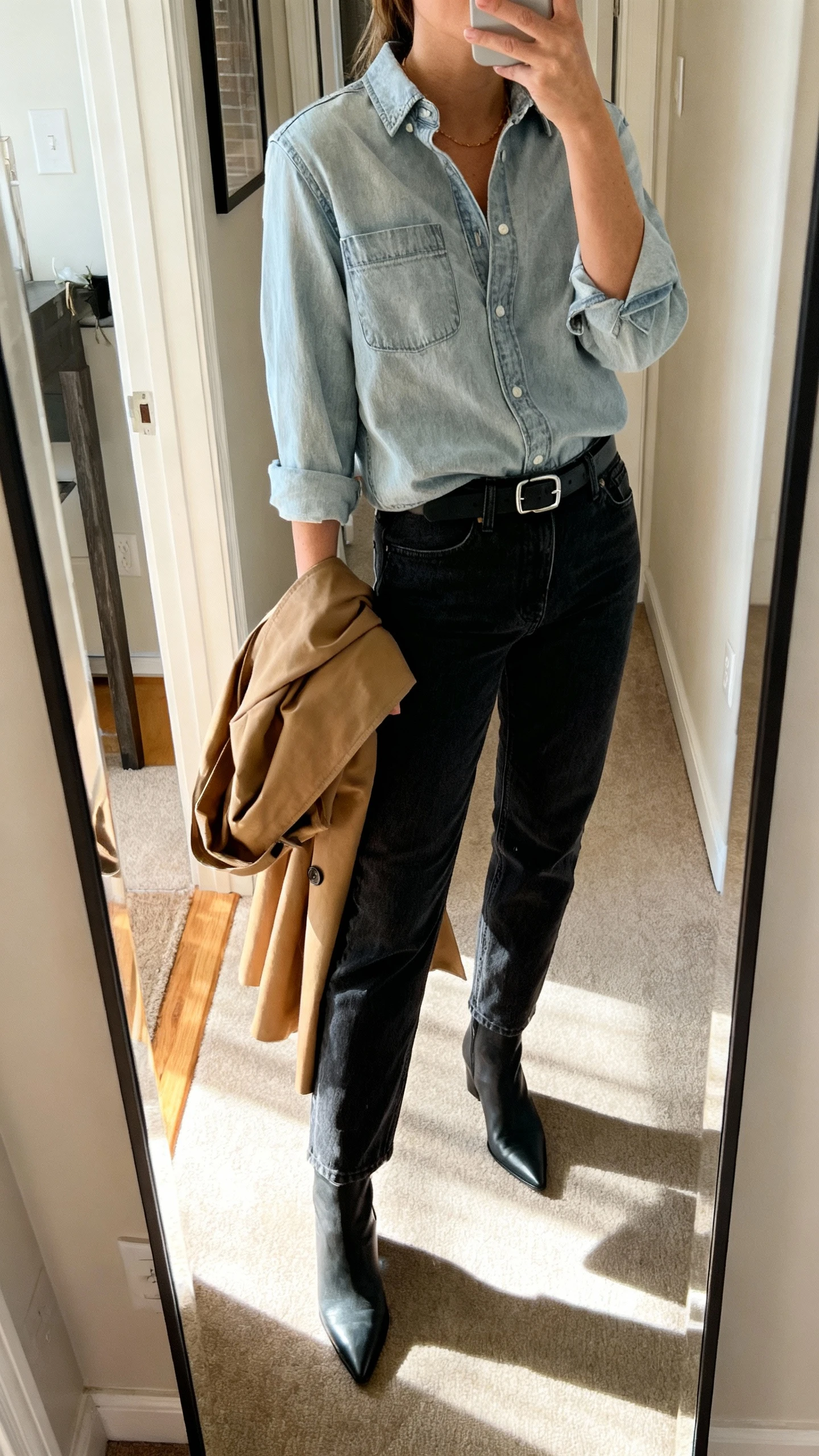 iPhone side-angle mirror selfie of a woman wearing a light-wash denim shirt tucked into dark-wash straight jeans, pointed ankle boots, minimalist belt, and a tan trench draped over the arm, face not visible, cropped at neck, entryway full-length mirror, indoor afternoon light, iPhone photo quality.