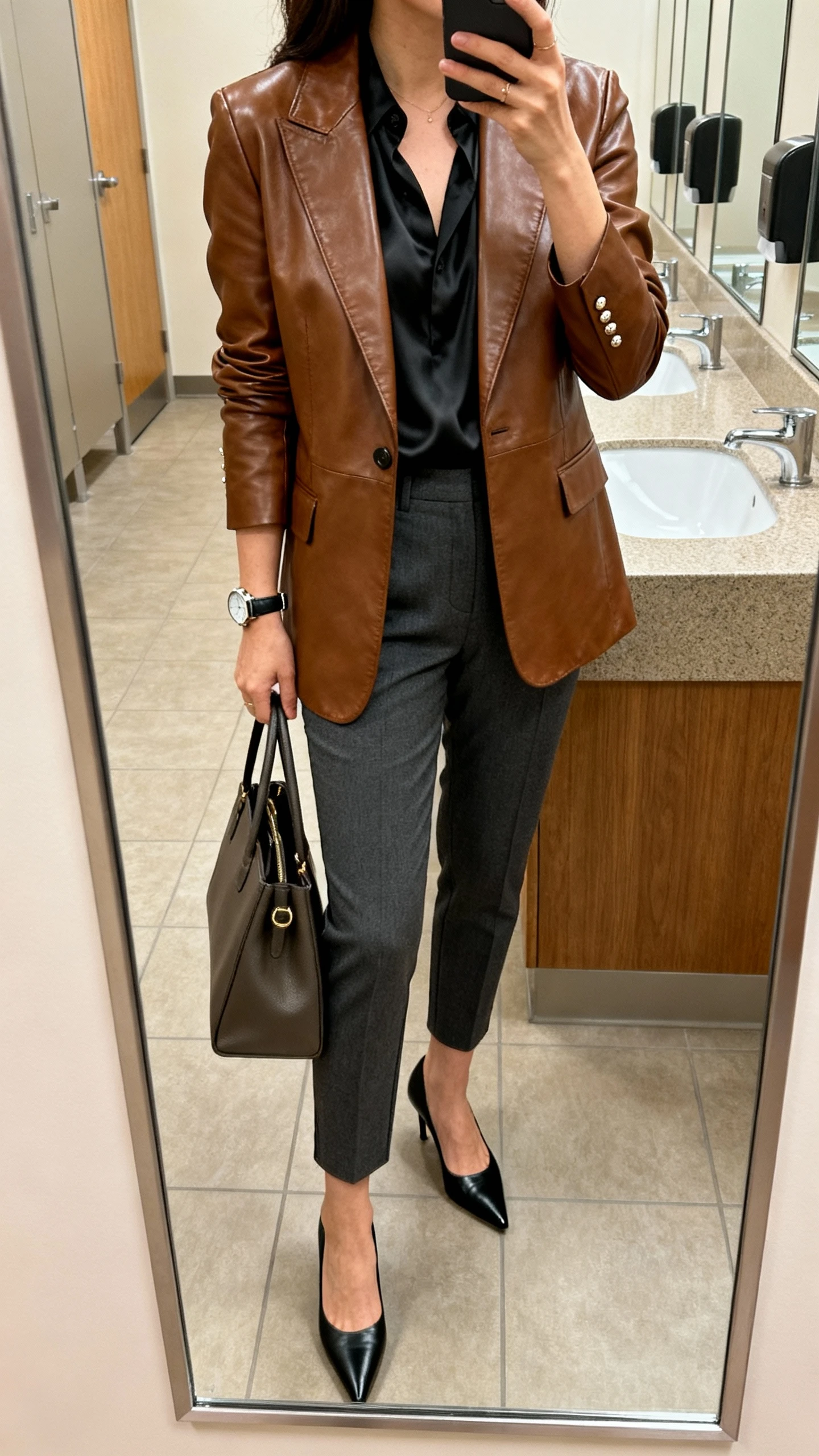 iPhone side-angle mirror selfie of a woman wearing a tailored brown leather blazer over a black silk blouse, charcoal tapered trousers, pointed black pumps, structured tote, watch and subtle studs, face not visible, office restroom mirror, warm indoor lighting, iPhone photo quality