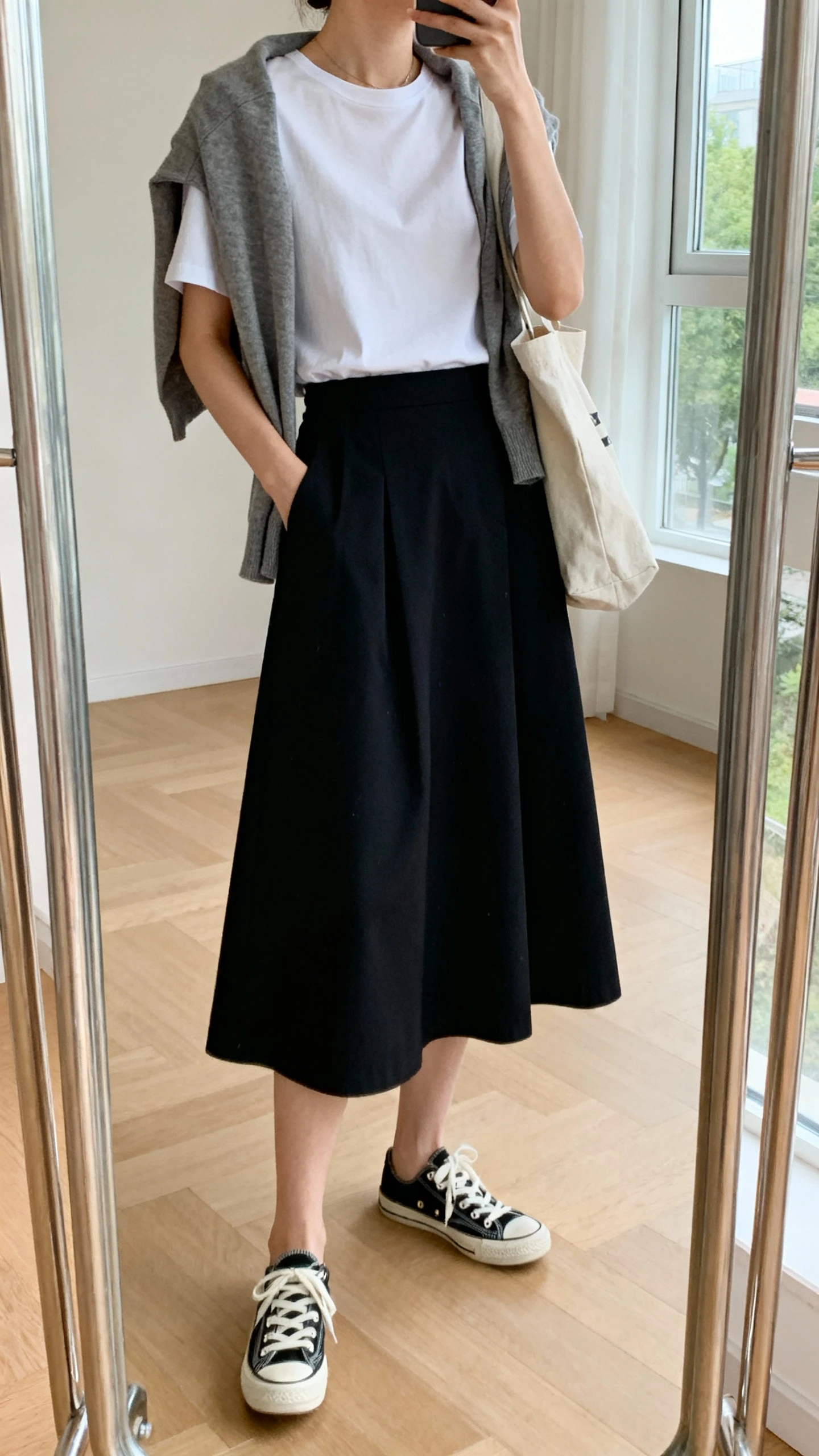iPhone side-angle mirror selfie of a woman wearing a midi A-line skirt with a tucked-in relaxed tee, classic lace-up sneakers, lightweight cardigan draped over shoulders, and a canvas tote, face not visible, cropped at neck, full-length mirror, soft window light, iPhone photo quality.
