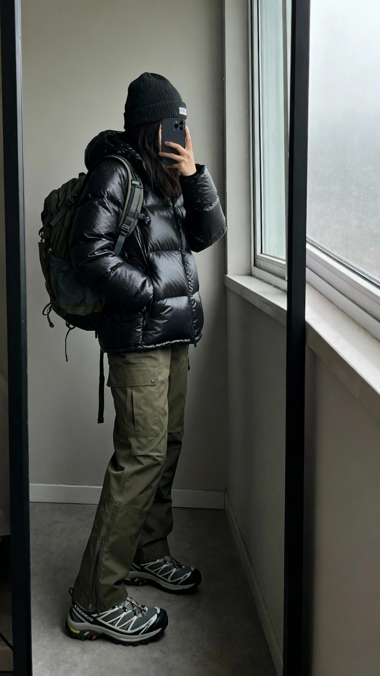 iPhone side-angle mirror selfie of a woman wearing a matte black puffer jacket, olive cargo pants, waterproof trail sneakers, beanie, and a practical backpack, face not visible, full-length mirror near a window with overcast natural light, iPhone photo quality.
