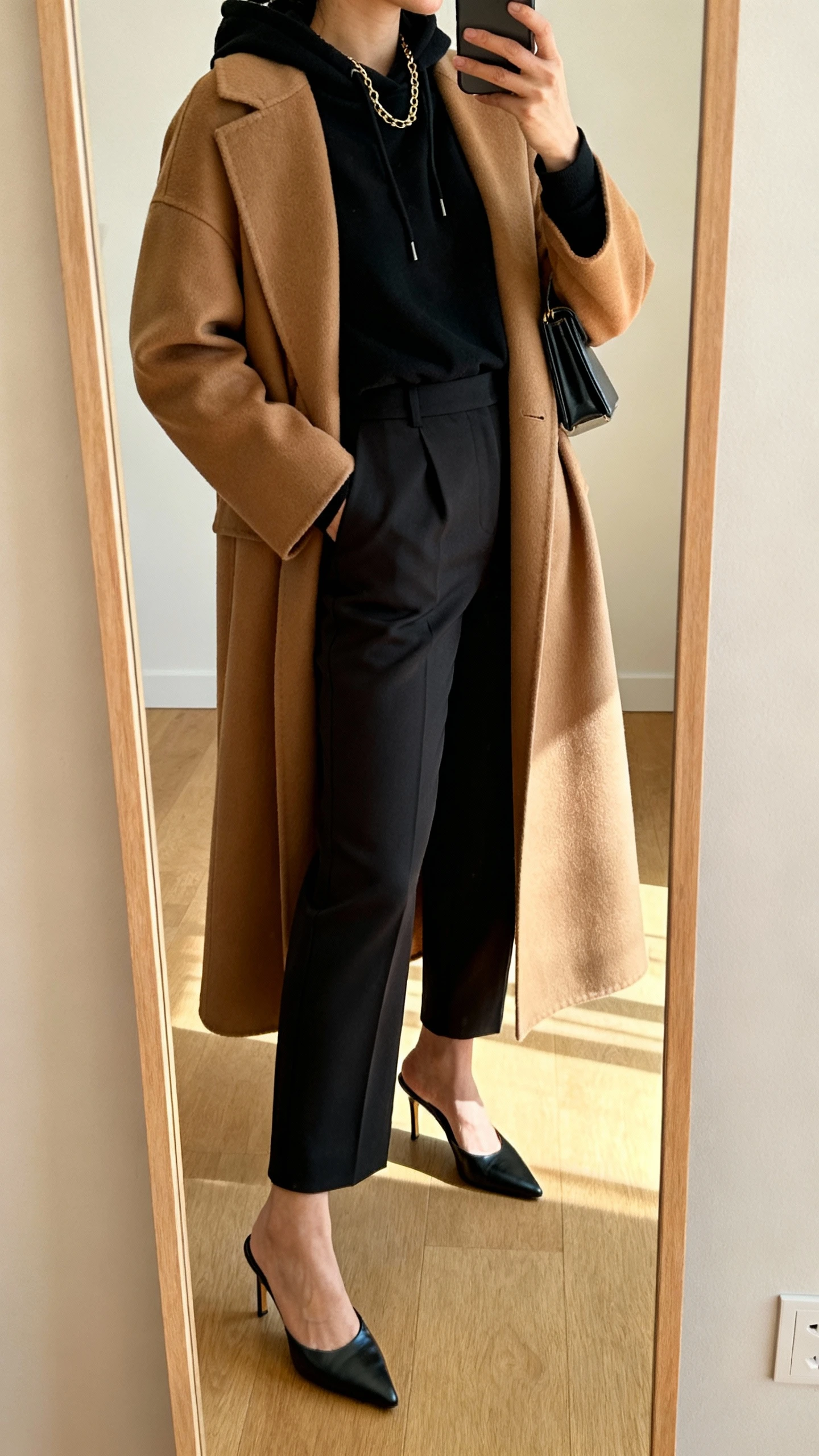 iPhone side-angle mirror selfie of a woman wearing a black hoodie with high-waisted dress pants, long statement coat in camel, sleek heeled mules, minimalist clutch, fine chain necklace, face not visible, full-length mirror, warm indoor lighting, iPhone photo quality.