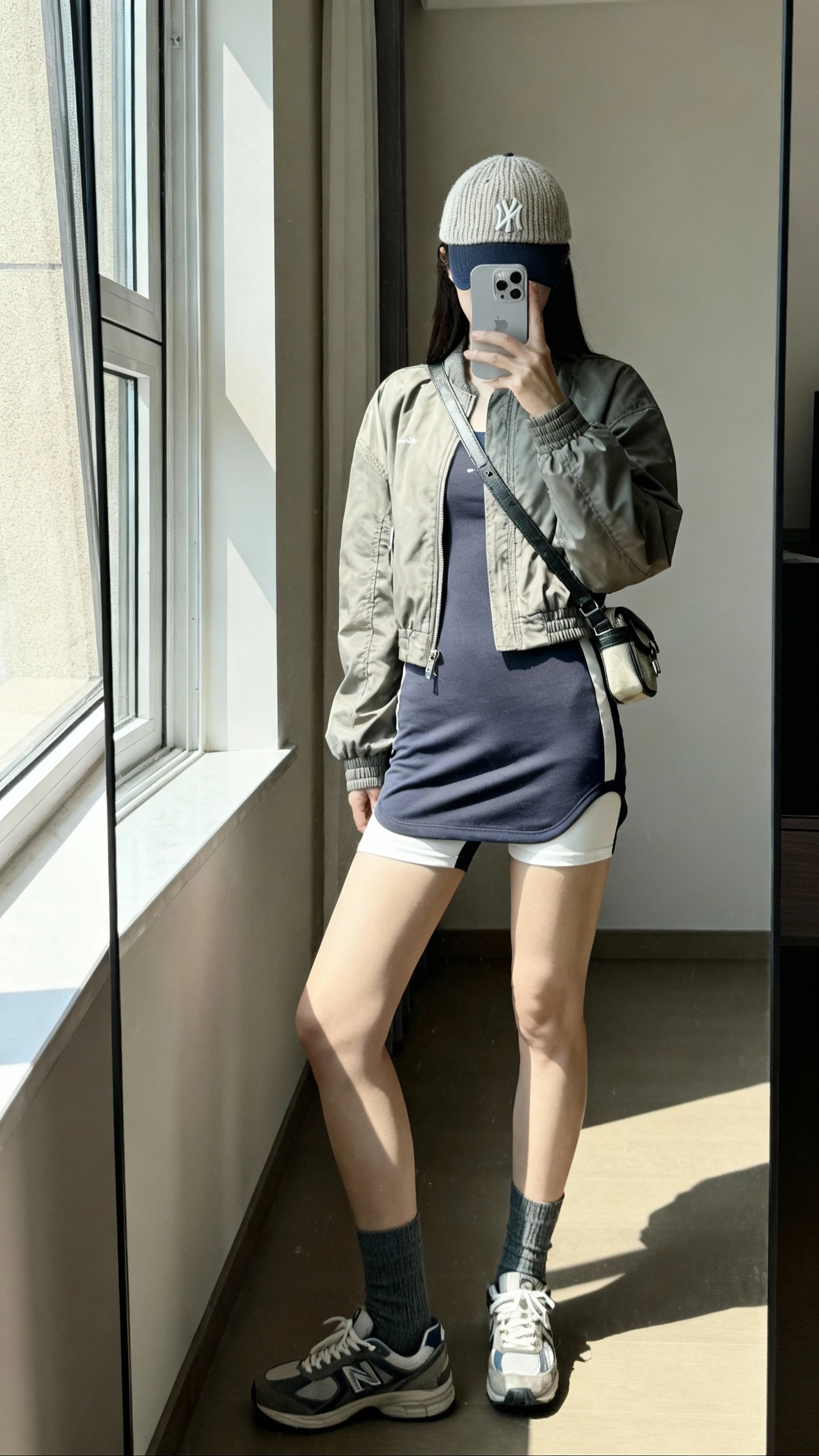 iPhone side-angle mirror selfie of a woman wearing a sporty athleisure dress with built-in shorts, lightweight bomber, knit baseball cap, ankle socks, retro trainers, crossbody belt bag, face not visible, full-length mirror near a window with bright natural light, iPhone photo quality.
