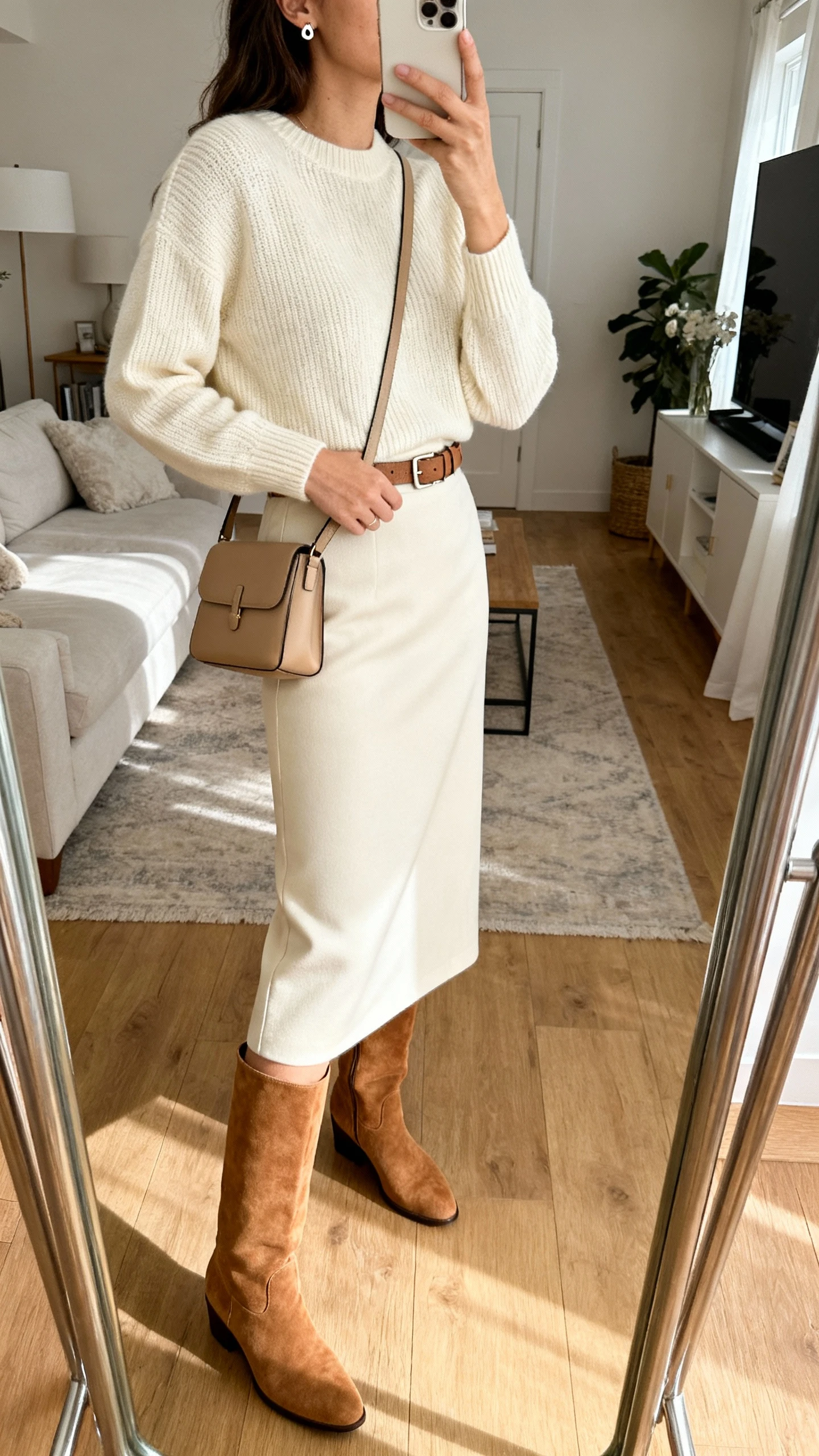 iPhone side-angle mirror selfie of a woman wearing monochrome suede knee-high boots, sleek midi skirt, fine-gauge knit sweater, minimalist belt, structured crossbody, and stud earrings, all in tonal cream and camel, face not visible, living room full-length mirror with natural morning light, iPhone photo quality.
