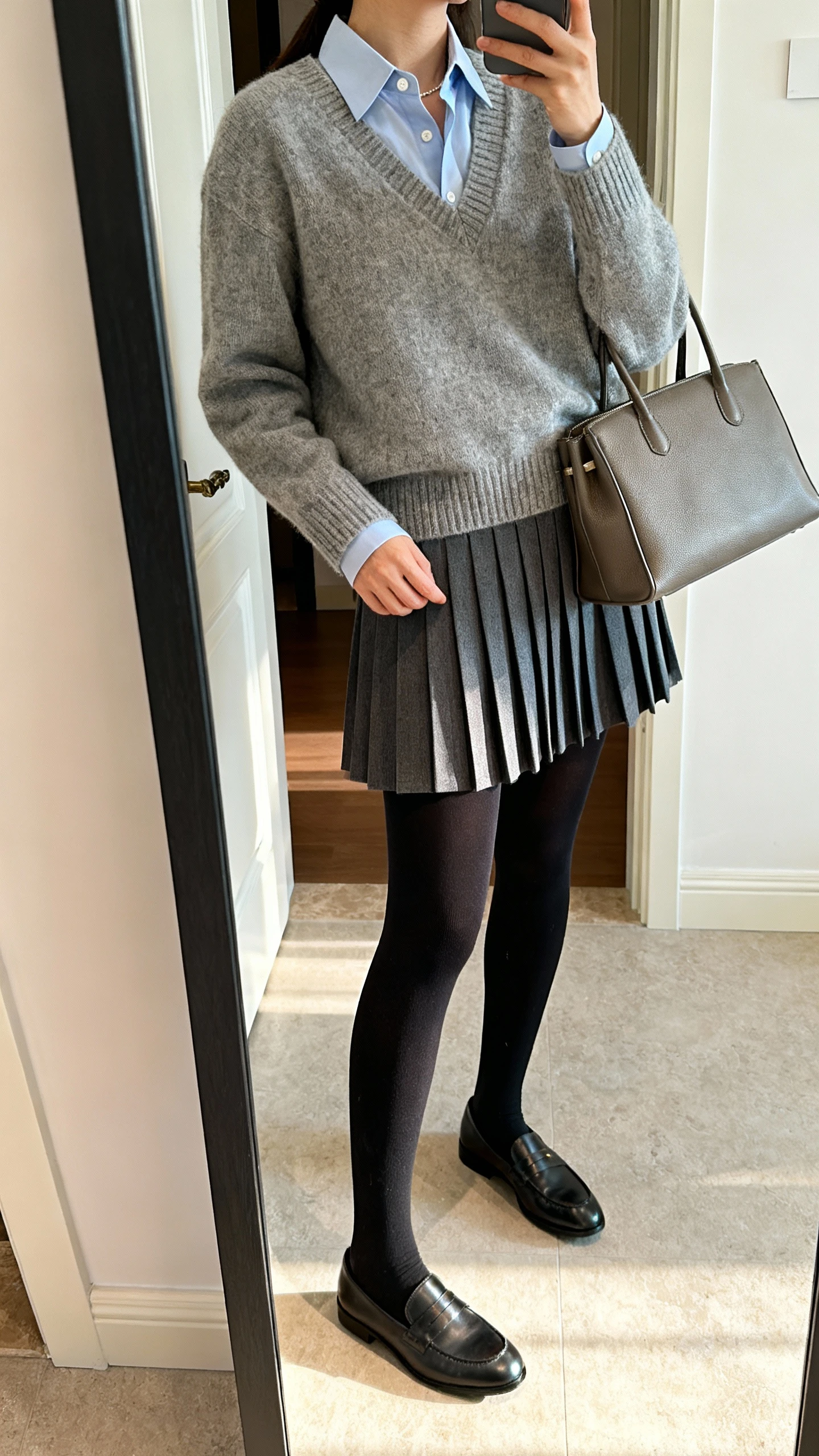 iPhone side-angle mirror selfie of a woman wearing a preppy pleated skirt, thermal black tights, button-up shirt under a V-neck sweater, loafers, and a structured tote, face not visible, cropped at neck, entryway full-length mirror, indoor daylight, iPhone photo quality.