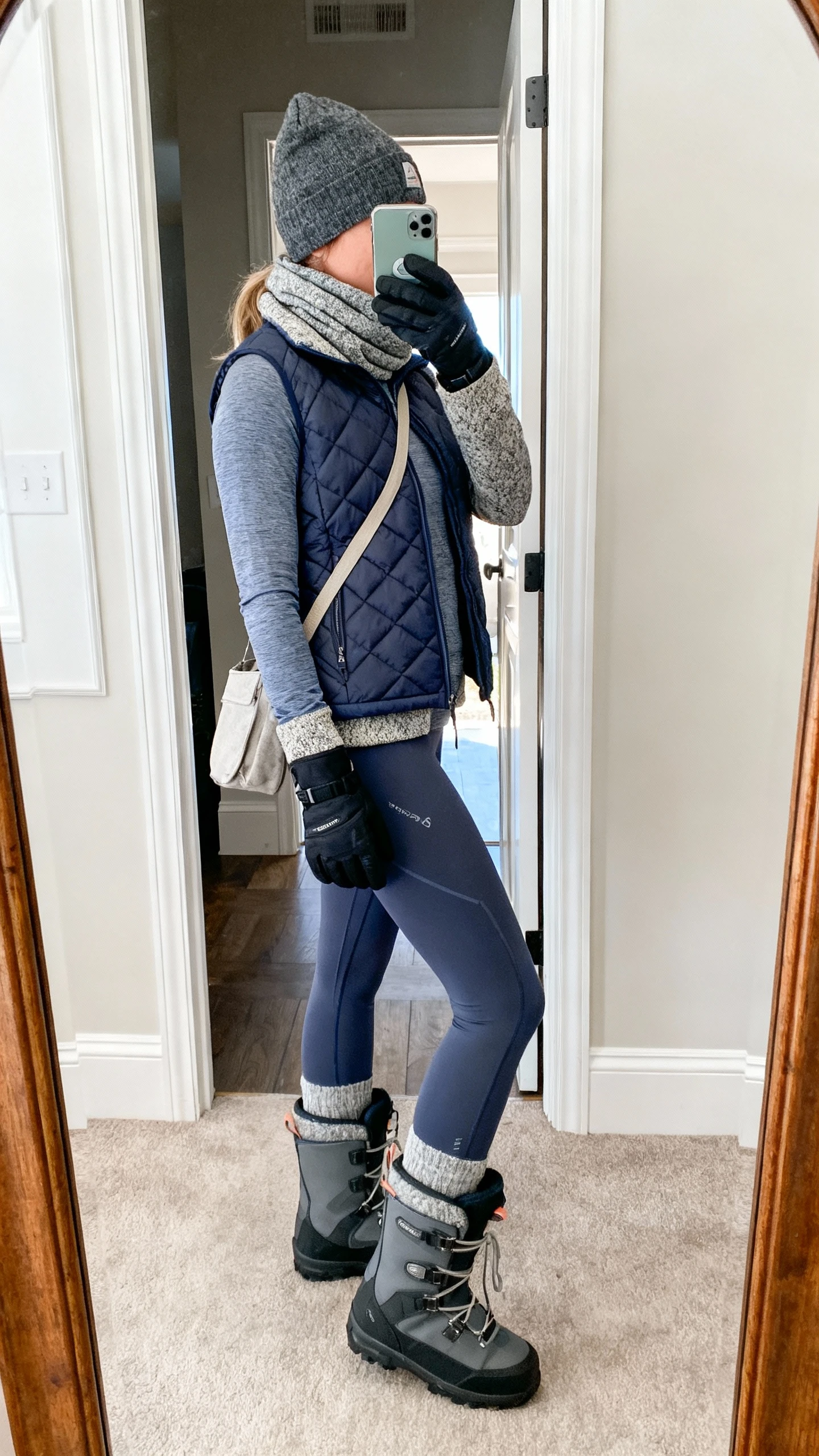 iPhone side-angle mirror selfie of a woman wearing a moisture-wicking base layer set (long-sleeve top and leggings), quilted insulated vest, wool-blend shacket, waterproof insulated snow boots, thermal beanie, neck gaiter, tech gloves, and a casual crossbody, face not visible, entryway full-length mirror, daytime indoor light, iPhone photo quality.