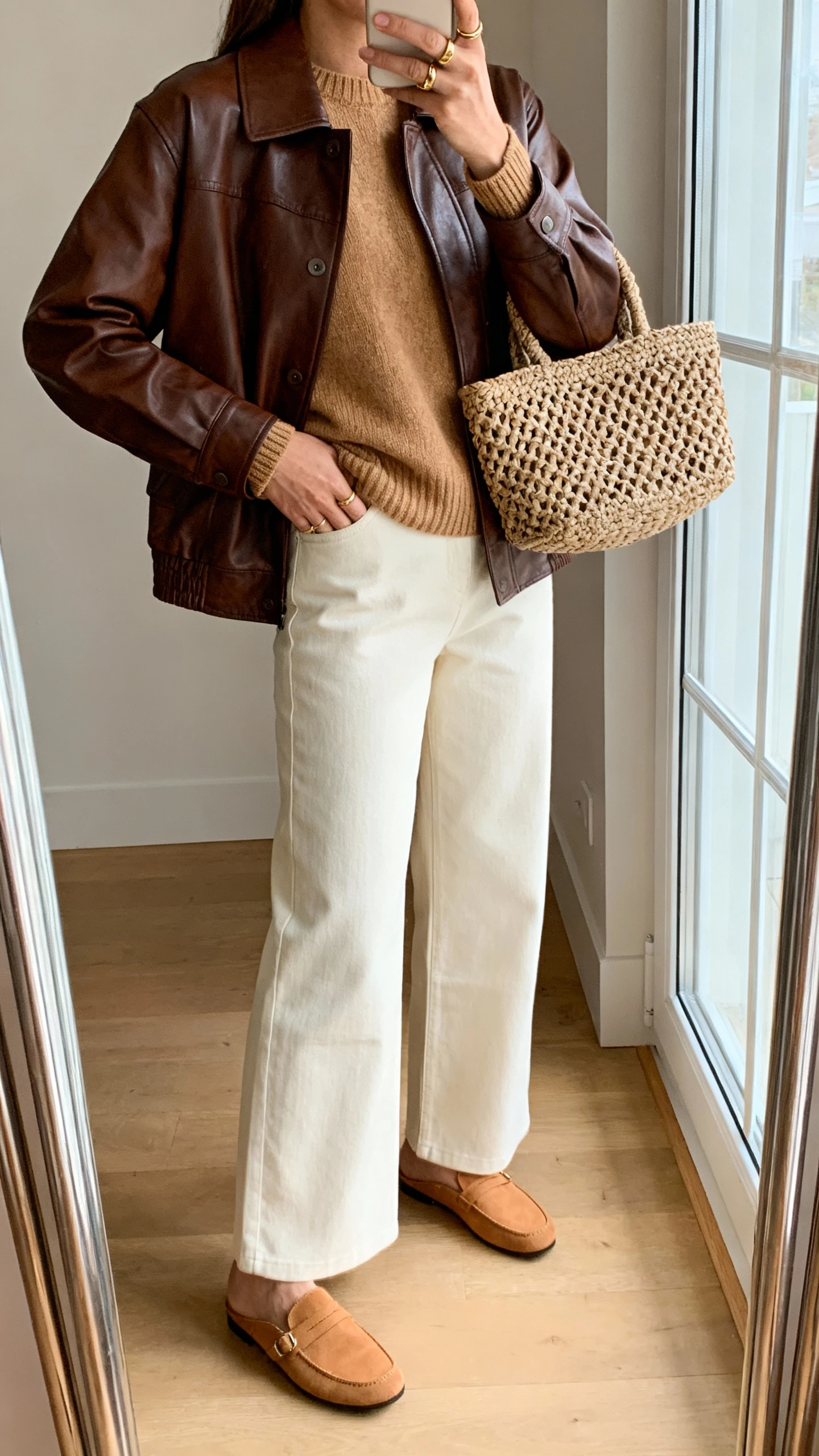 iPhone side-angle selfie of a woman wearing a brown leather jacket with cream wide-leg trousers, camel knit sweater, tan loafers, and a textured woven tote, plus simple gold rings, face not visible, full-length mirror near window, warm indoor light, iPhone photo quality.