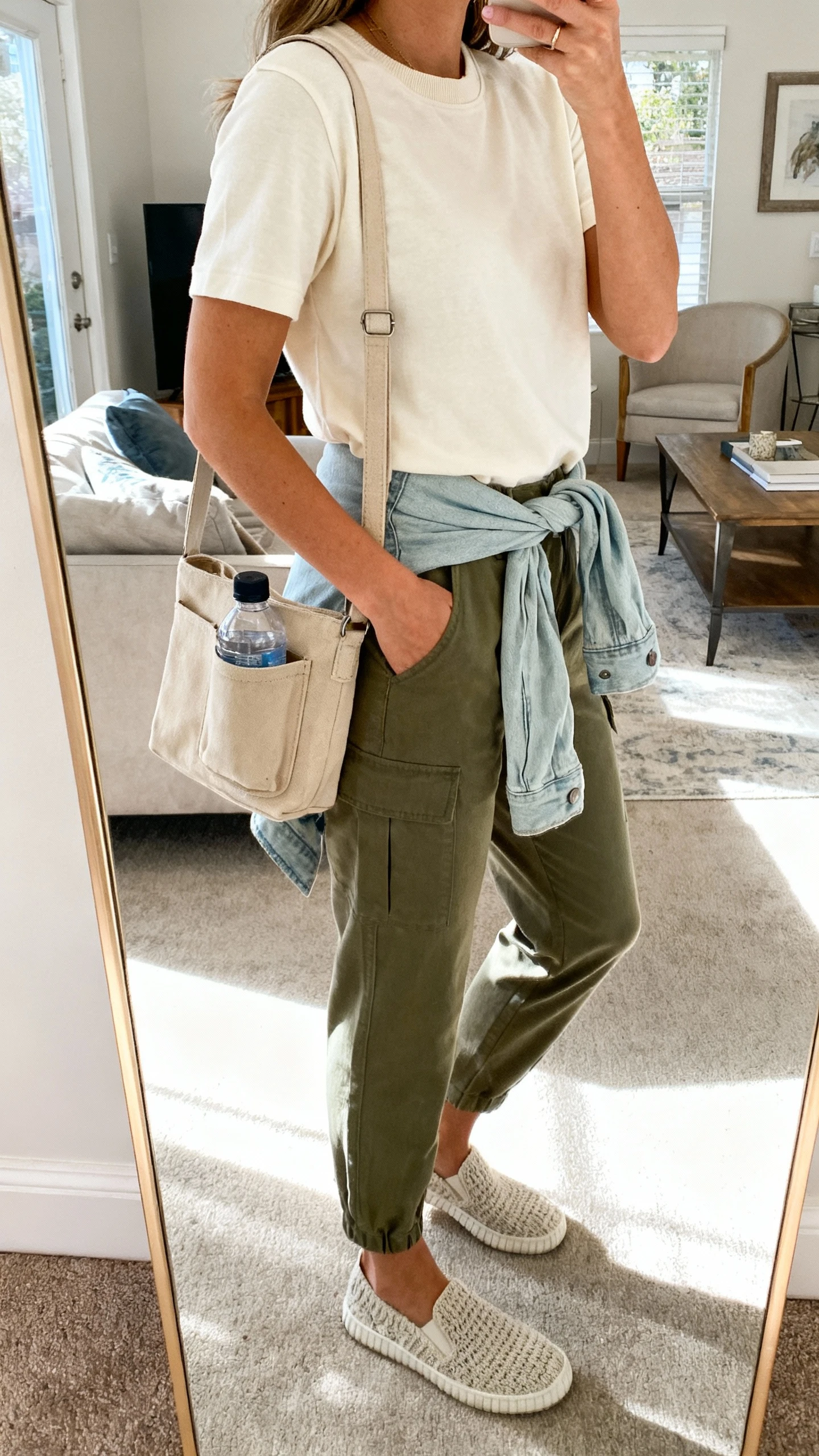 iPhone side-angle selfie of a woman wearing a soft cream crewneck tee, relaxed olive utility pants, comfortable knit slip-on sneakers, a sand-colored canvas crossbody with water bottle pocket, and a lightweight denim jacket tied at the waist, face not visible, living-room mirror, bright natural daylight, iPhone photo quality