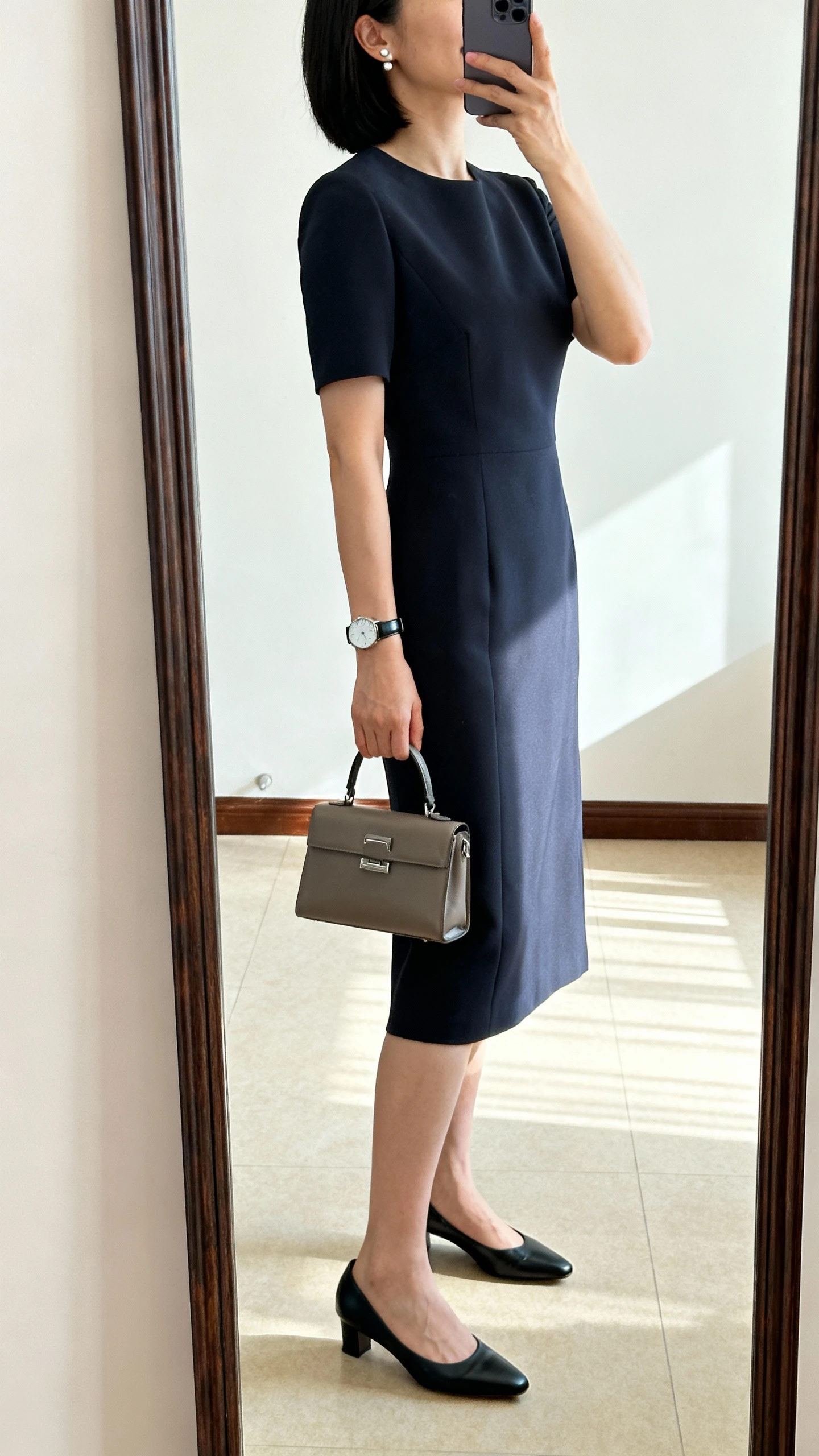 iPhone side-angle selfie of a woman wearing a tailored midi sheath dress with low kitten heels, a structured mini tote, stud earrings, and a sleek watch, face not visible, full-length mirror, bright indoor lighting, iPhone photo quality.