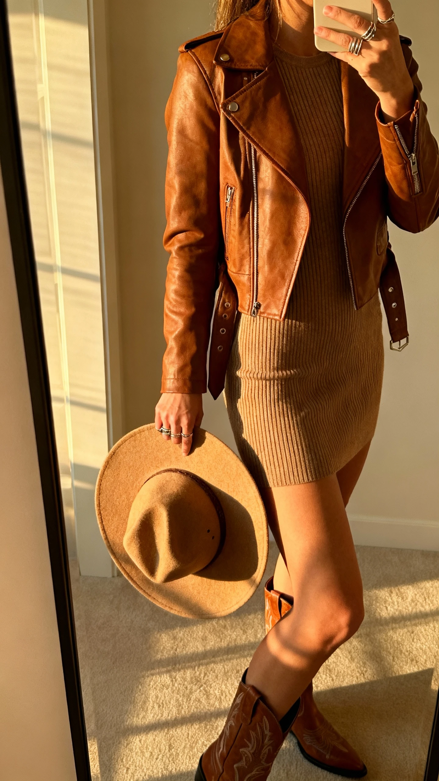 iPhone side-view mirror selfie of a woman wearing a brown leather jacket over a ribbed knit dress, Western-style ankle boots, felt rancher hat held at her side, layered rings, face not visible, full-length mirror, golden afternoon indoor light, iPhone photo quality