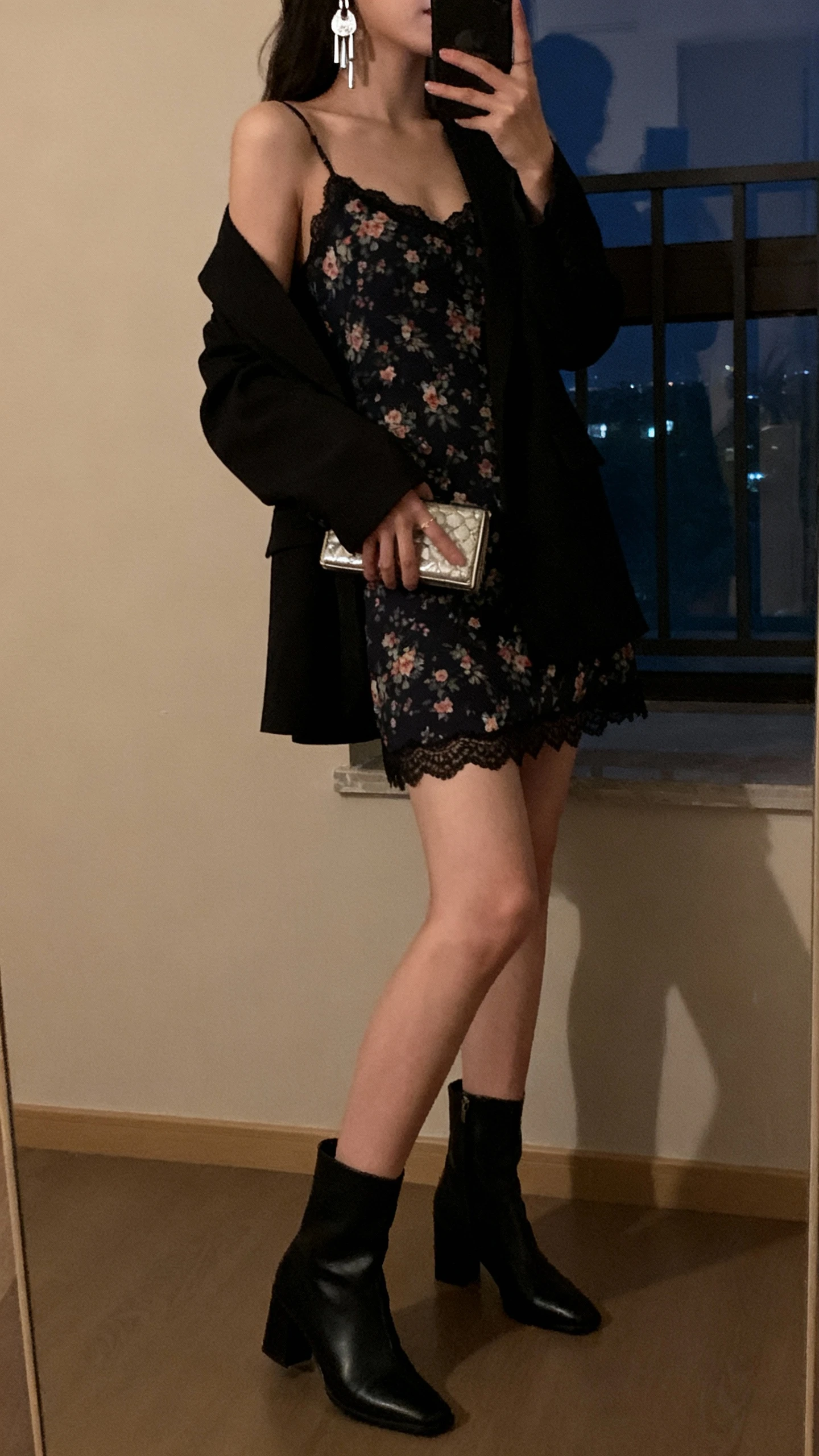 iPhone side-view selfie of a woman wearing a dark floral slip dress with lace trim, black heeled ankle boots, lightweight black blazer over shoulders, silver drop earrings, and a compact clutch; face not visible, cropped at shoulders, dim indoor evening light near window, iPhone photo quality.
