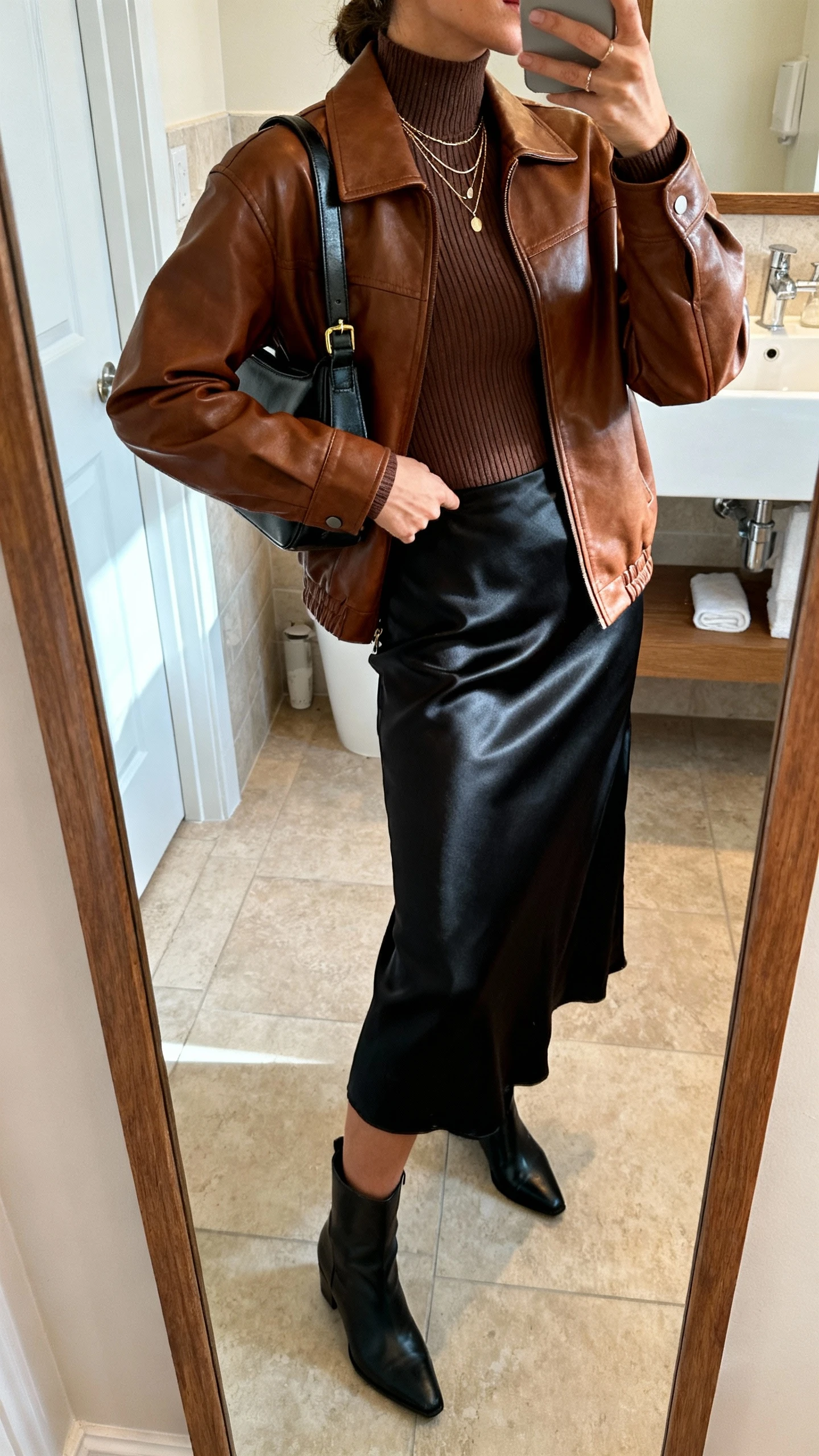 Side-angle iPhone mirror selfie of a woman wearing a brown leather jacket with a black satin midi skirt, fitted ribbed turtleneck, ankle boots, structured shoulder bag, and delicate layered necklaces, face not visible, bathroom full-length mirror, soft morning light, iPhone photo quality.