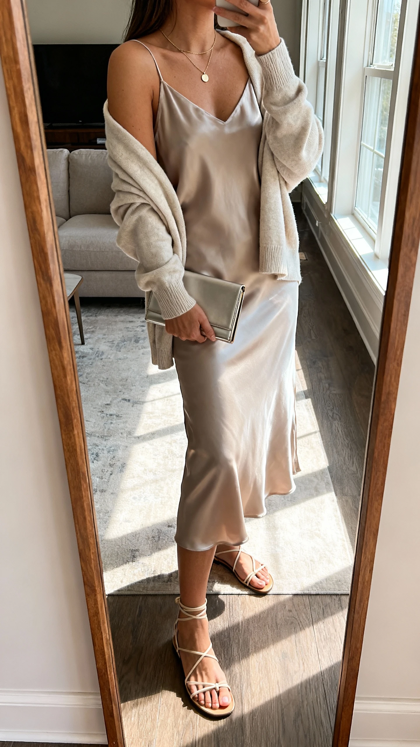 Side-angle iPhone mirror selfie of a woman wearing a slip dress and knit combo: silky midi slip dress with a lightweight crewneck cardigan draped over shoulders, strappy minimalist sandals, fine pendant necklace, slim clutch; face not visible, cropped at shoulders, full-length mirror in living room with natural window light, iPhone photo quality.