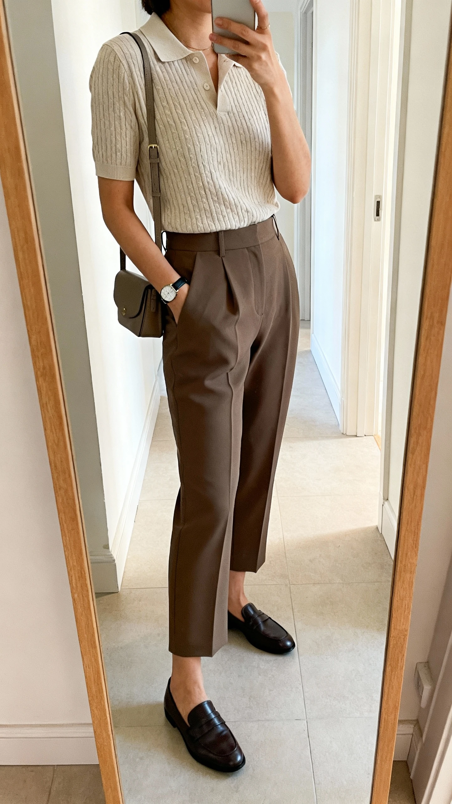 Side-angle iPhone mirror selfie of a woman wearing a short-sleeve knit polo tucked into tailored culottes, sleek loafers, slim watch, crossbody bag, face not visible, cropped at shoulders, full-length mirror in hallway, natural daylight, iPhone photo quality.