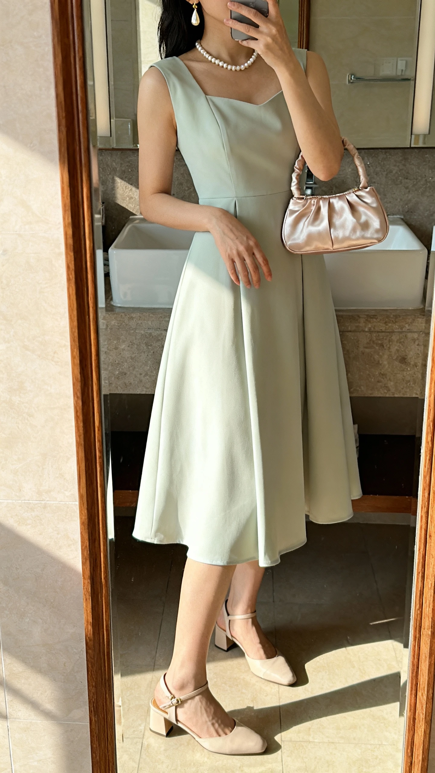 Side-angle iPhone mirror selfie of a woman wearing a tea-length A-line dress with elegant slingback heels, a pearl necklace, vintage-inspired drop earrings, and a satin top-handle bag, face not visible, cropped at shoulders, bathroom mirror, morning light, iPhone photo quality.