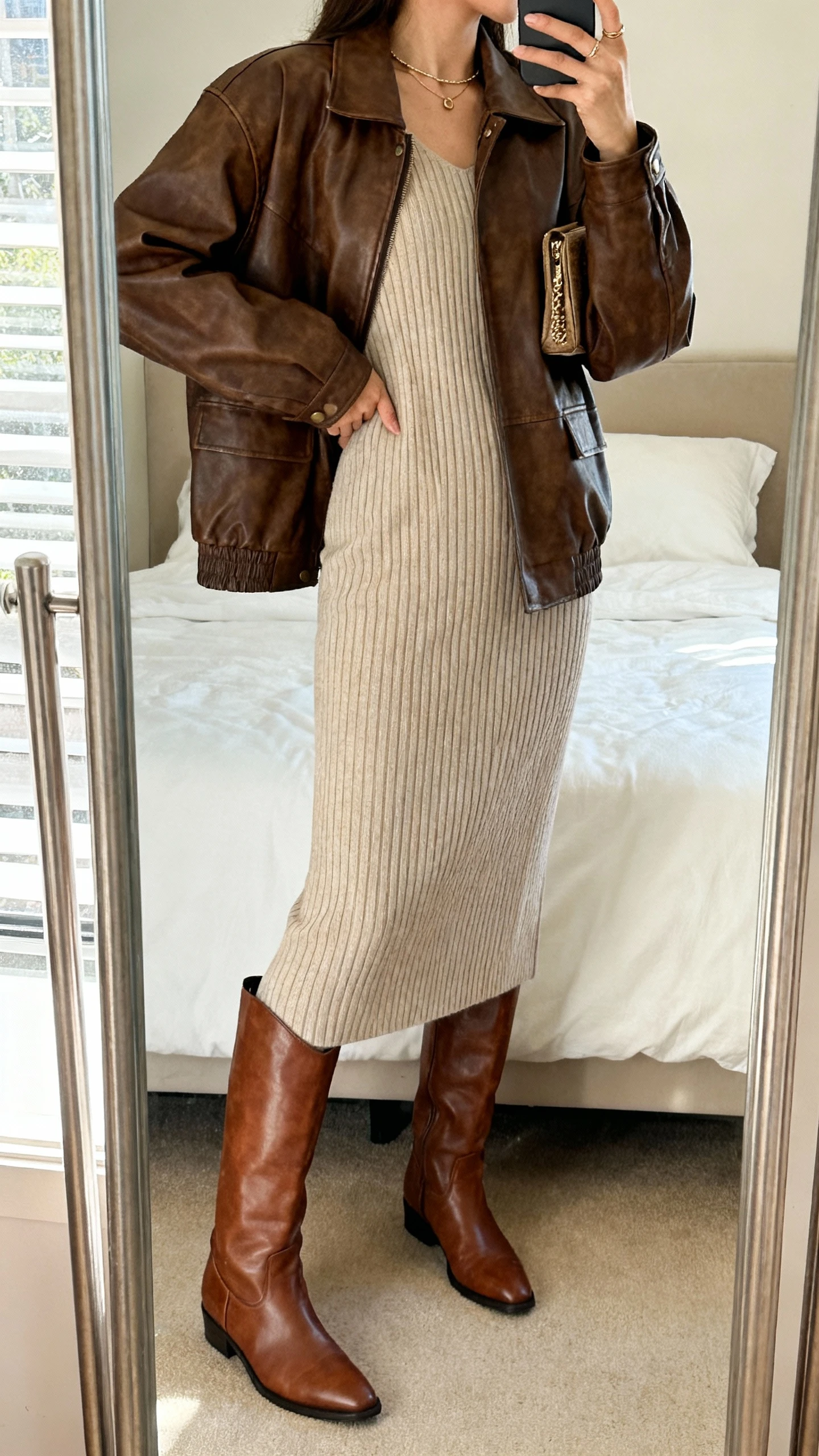 Side-angle iPhone mirror selfie of a woman wearing a ribbed knit midi dress and knee-high brown leather boots layered with a brown leather jacket, minimal jewelry and a small clutch, face not visible, cropped at shoulders, bedroom mirror, natural daylight, iPhone photo quality