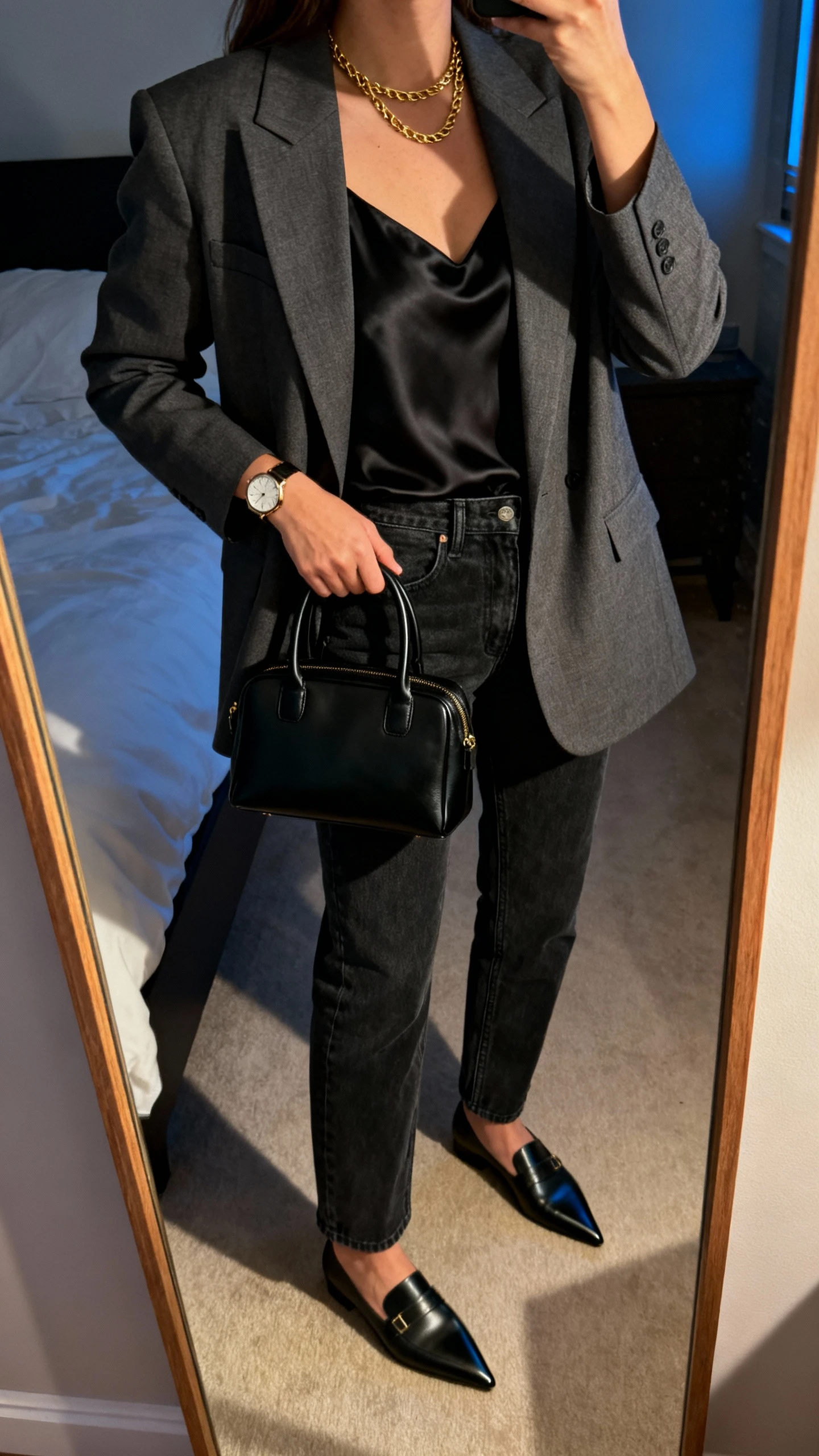 Side-angle iPhone mirror selfie of a woman wearing a tailored charcoal blazer over a black silk camisole, straight-leg dark jeans, pointed-toe loafers, structured black top-handle bag, slim gold chain and watch, face not visible, cropped at shoulders, bedroom mirror with indoor evening light, iPhone photo quality