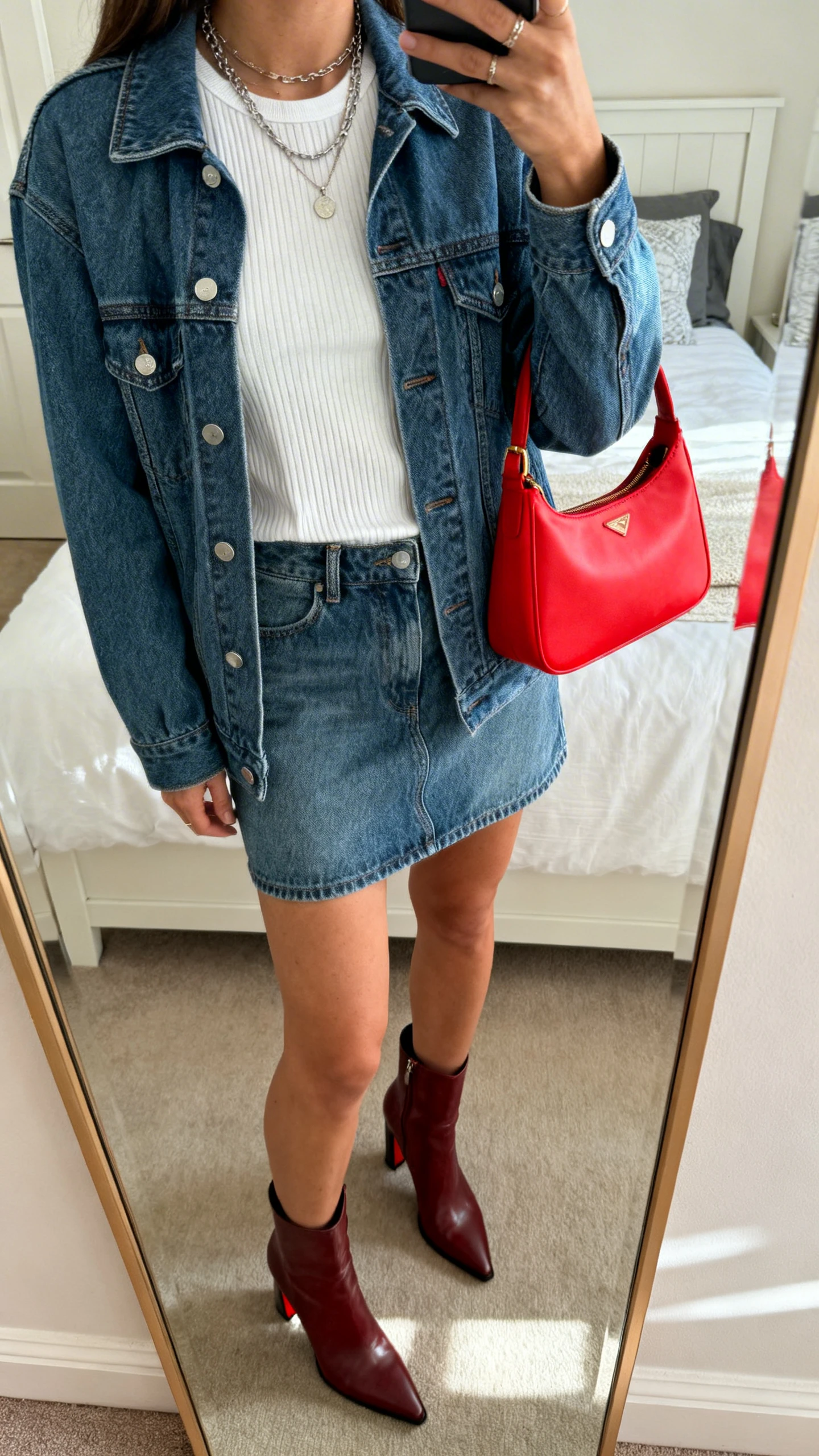 Cropped-at-shoulders iPhone mirror selfie of a woman wearing a denim jacket over a denim mini skirt, white ribbed tee, bright red bag for a pop of color, heeled ankle boots, and layered necklaces, face not visible, bedroom mirror, natural daylight, iPhone photo quality.