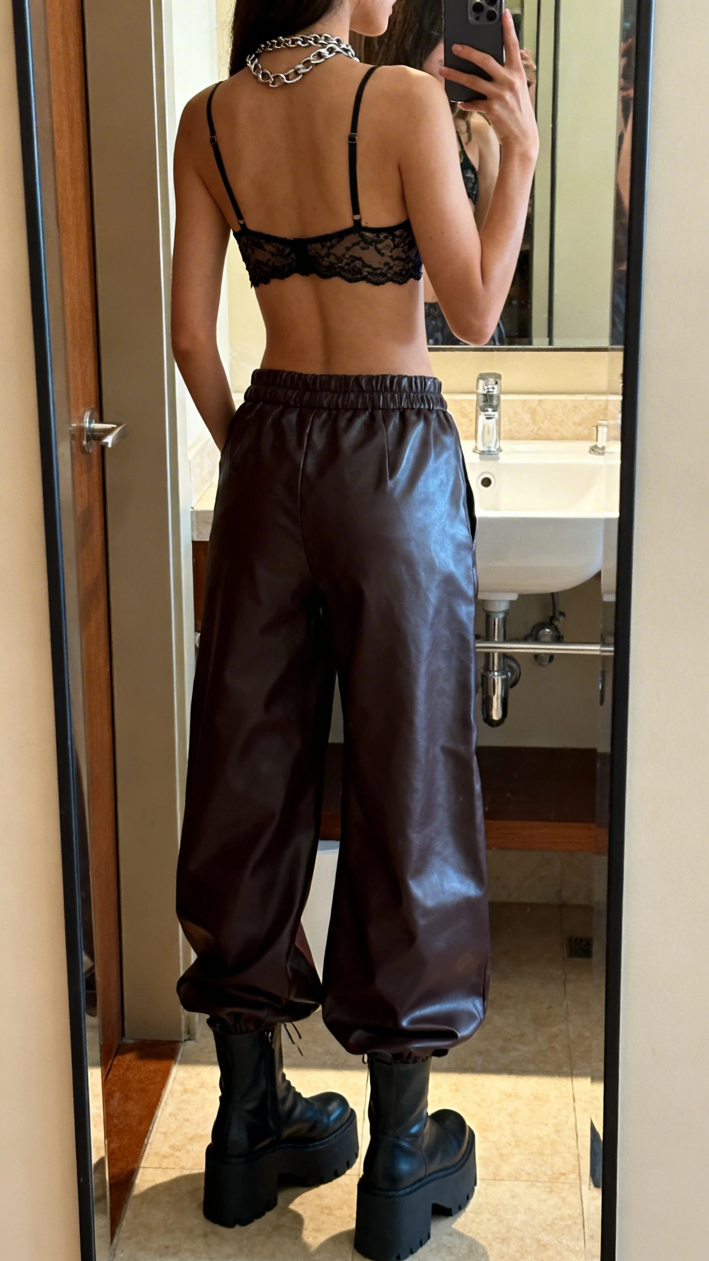 iPhone back-view mirror selfie of a woman wearing wide-leg faux leather pants, cropped lace bralette, chunky platform boots, layered chain necklace, face not visible, bathroom full-length mirror, warm indoor lighting, iPhone photo quality.