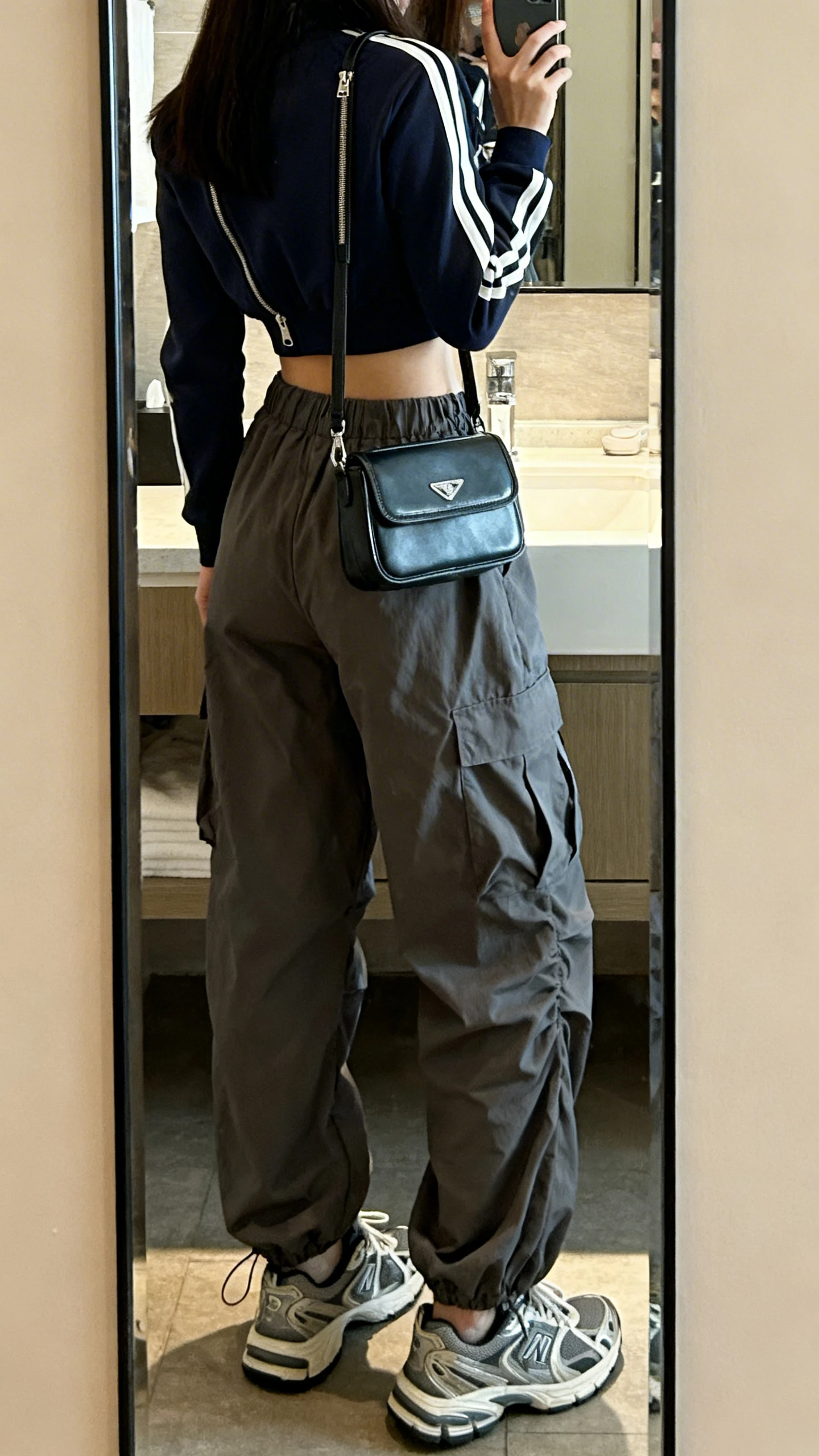 iPhone back-view mirror selfie of a woman wearing parachute cargo pants and a sporty zip-up crop top, chunky trainers, mini shoulder bag, face not visible, bathroom mirror, soft indoor lighting, iPhone photo quality.