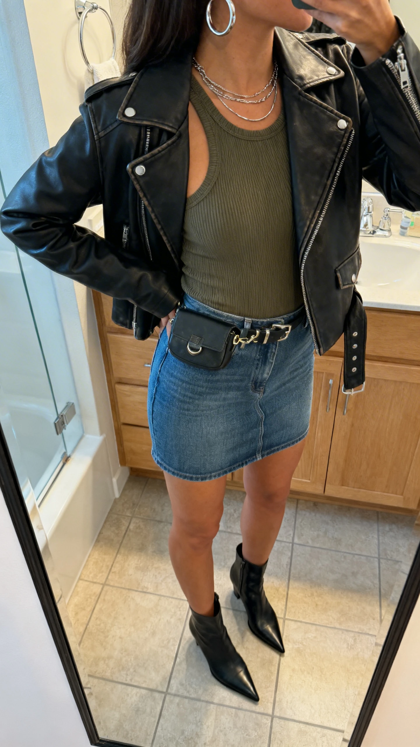 iPhone back-view mirror shot of a woman wearing a leather jacket over a fitted denim mini skirt and ribbed tank, with pointed ankle boots, layered silver hoops, and a small belt bag, face not visible, bathroom mirror, soft indoor lighting, iPhone photo quality.