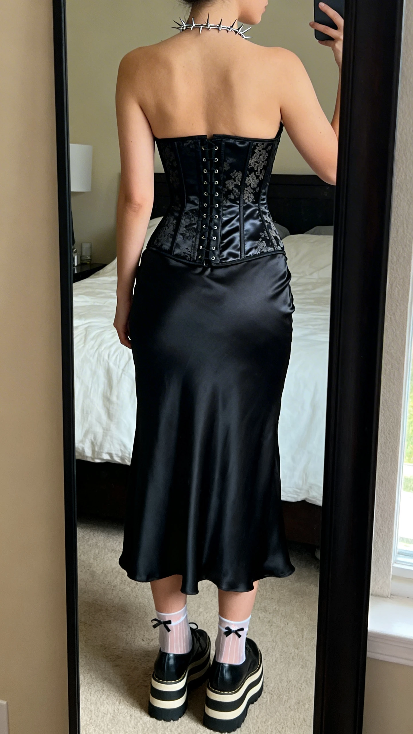 iPhone back-view mirror shot of a woman wearing a bias-cut black slip dress (midi/tea length) cinched with a black satin/brocade underbust corset, platform Mary Janes or stacked creepers, sheer ankle socks with tiny bows, and a sleek spiked/barbed-wire-inspired necklace, face not visible, bedroom mirror, warm indoor lighting, iPhone photo quality.