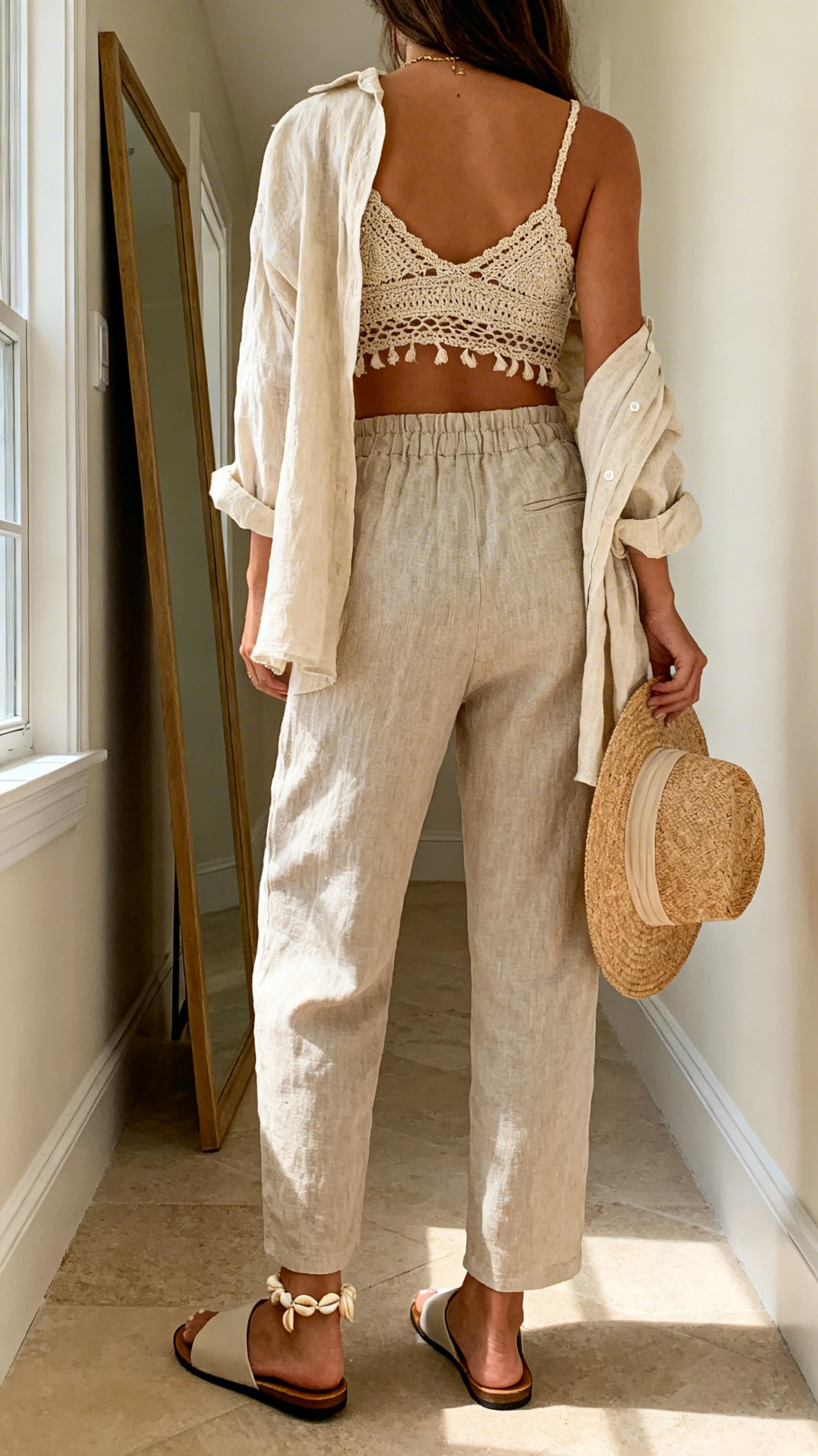 iPhone back view of a woman wearing a desert goddess crochet bralette with a matching linen set (open linen shirt and high-waist linen trousers), flat slide sandals, shell anklet, and a straw hat held at her side, face not visible, hallway full-length mirror, soft afternoon window light, iPhone photo quality.