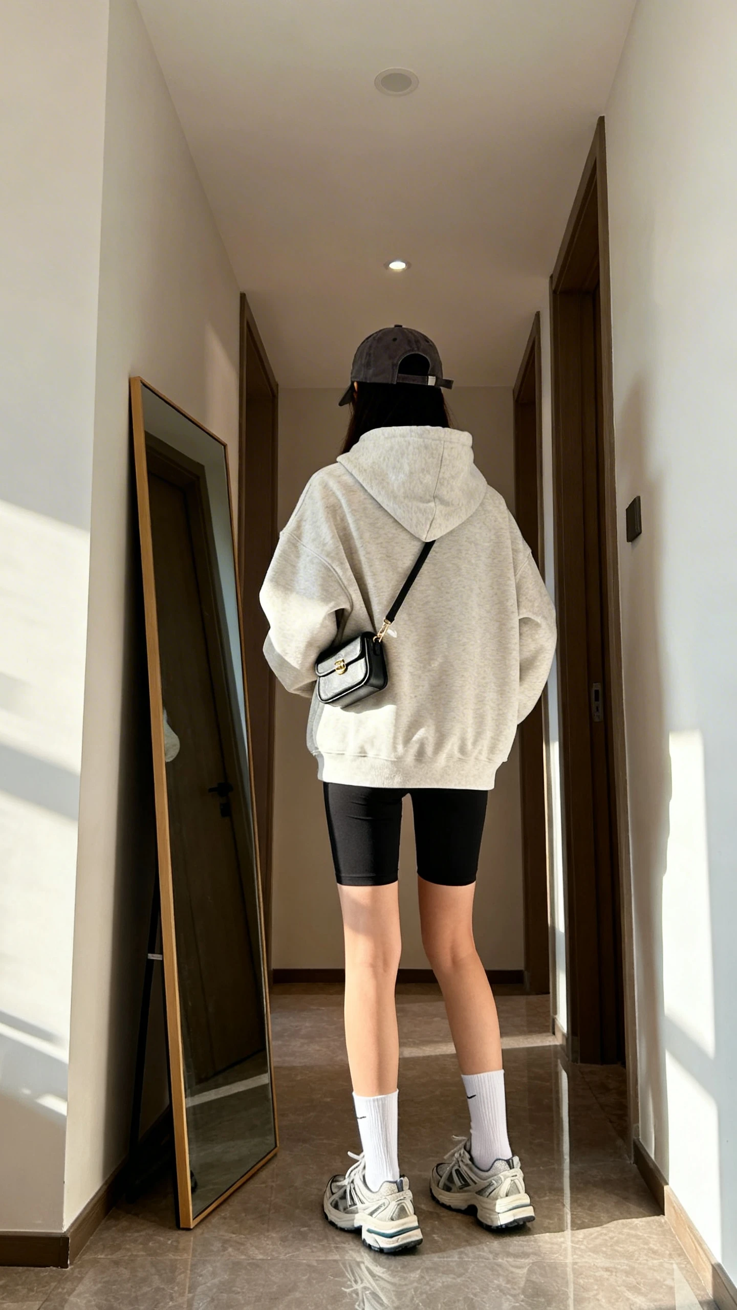 iPhone back view of a woman wearing an oversized hoodie, black biker shorts, white crew socks, dad sneakers, a baseball cap, and a tiny shoulder bag, face not visible, hallway full-length mirror, morning light, iPhone photo quality.