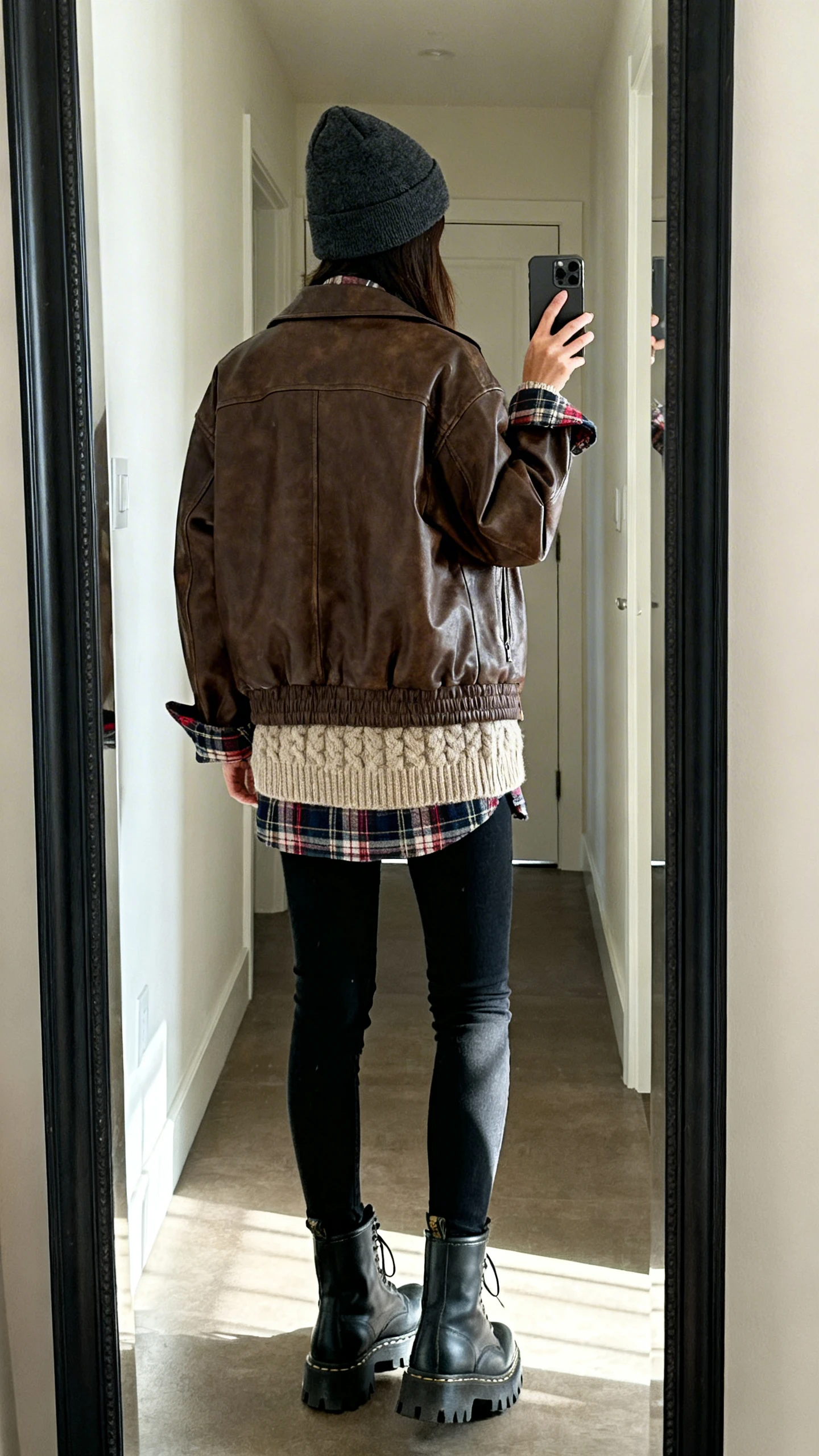 iPhone back-view selfie in a full-length mirror of a woman wearing a slightly oversized brown leather jacket, chunky knit sweater in oatmeal, plaid flannel peeking at cuffs and collar, black skinny jeans, lug-sole combat boots, and a dark gray beanie, face not visible, hallway mirror with morning light, iPhone photo quality.