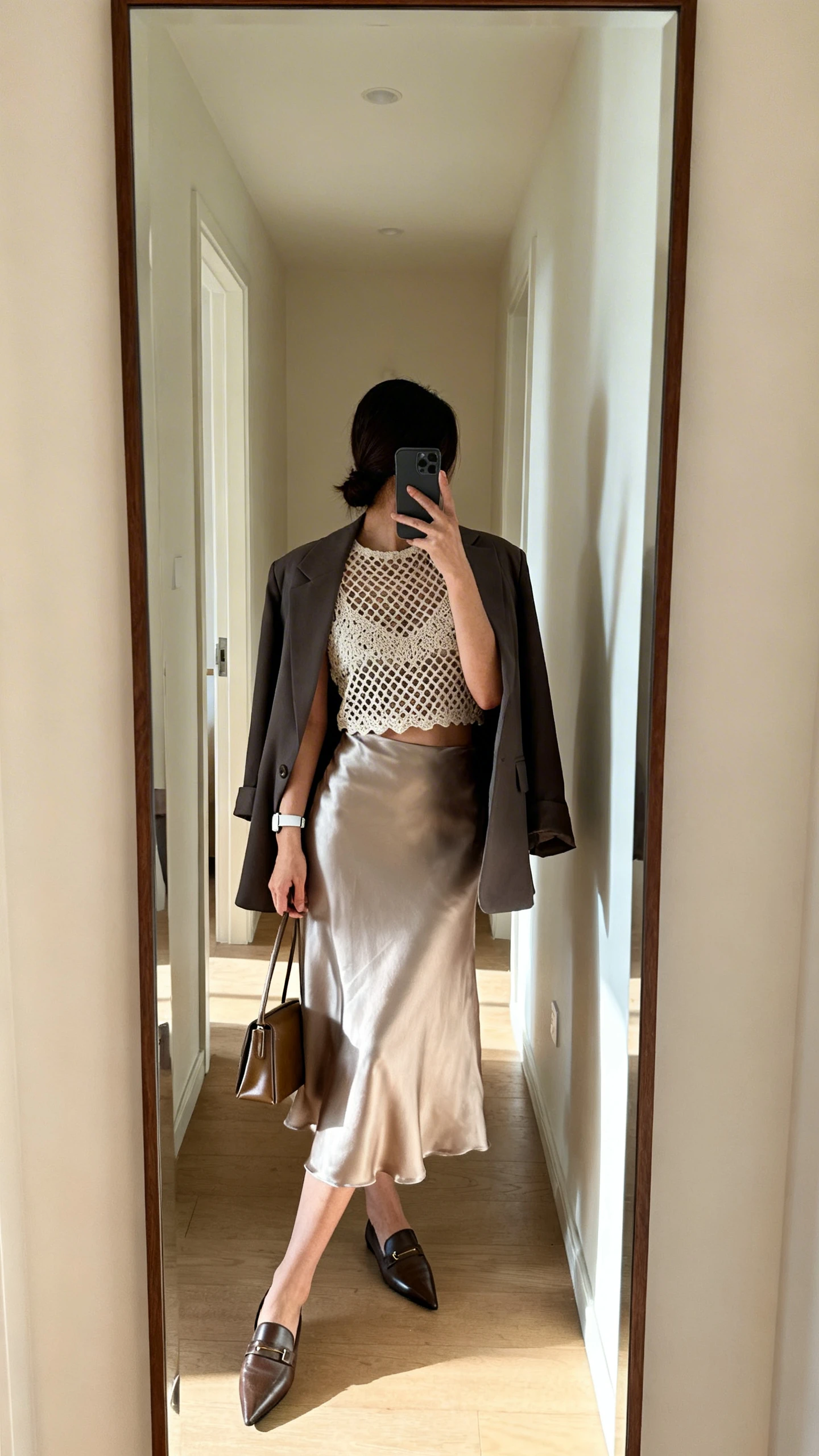 iPhone back-view selfie of a woman wearing a crochet mesh top tucked into a satin slip skirt, structured blazer draped over shoulders, pointed-toe loafers, slim leather tote, and a minimalist watch; face not visible, hallway full-length mirror, morning light, iPhone photo quality.