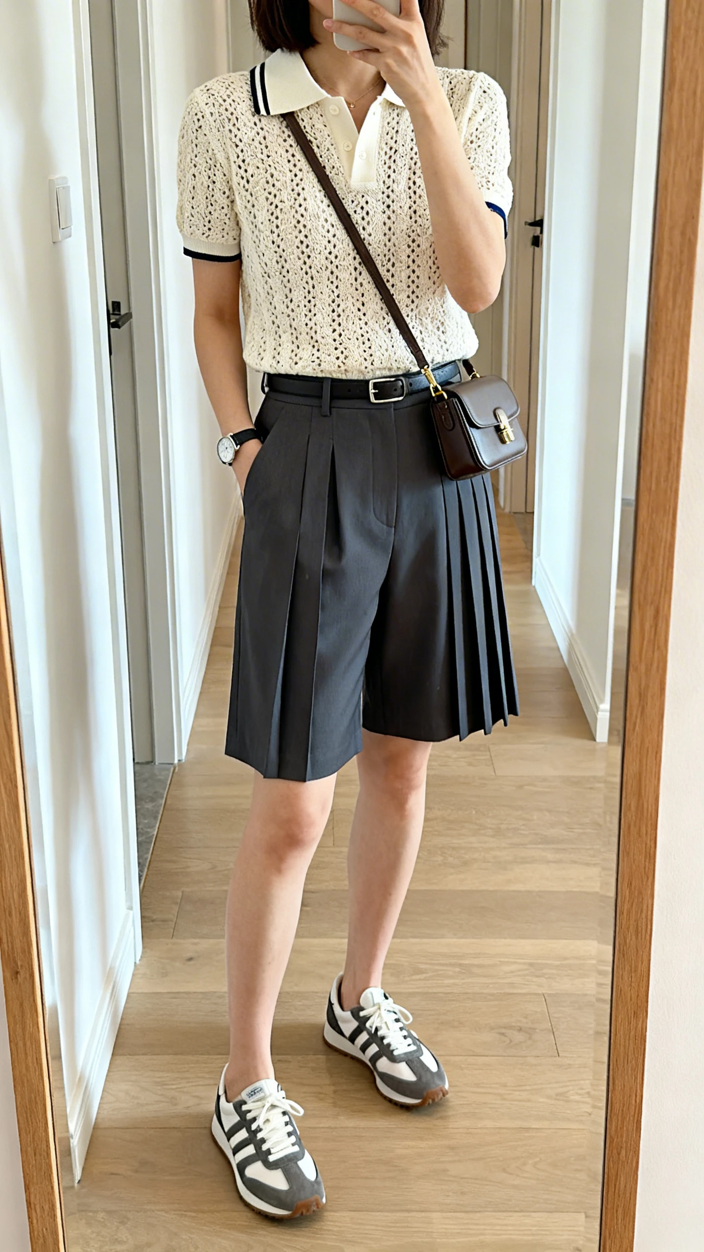 iPhone full-length mirror selfie of a woman wearing a sporty crochet polo tucked into tailored pleated shorts, sleek low-profile sneakers, minimalist watch, and a compact crossbody bag; face not visible, cropped at neck, hallway mirror, bright indoor lighting, iPhone photo quality.