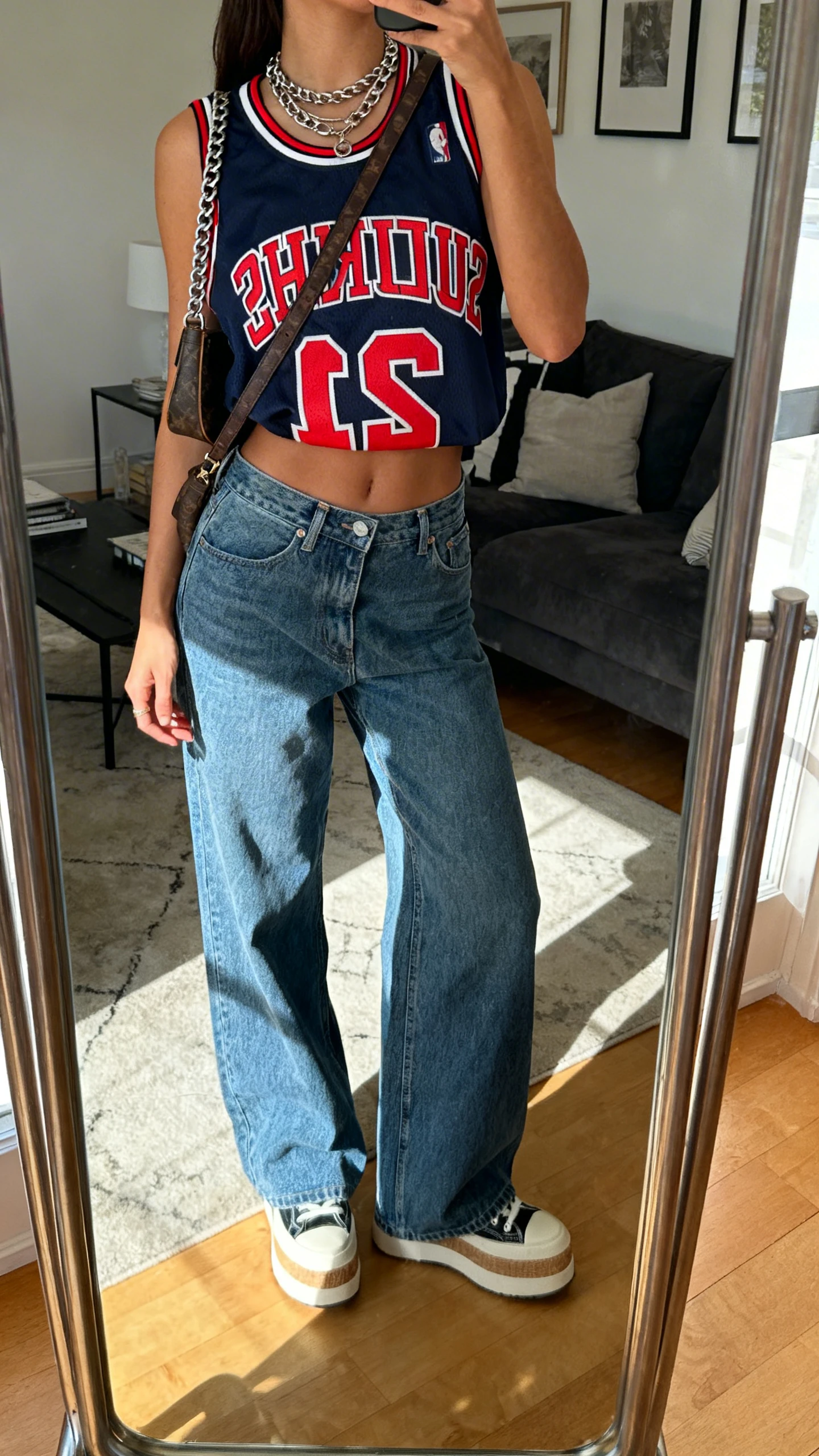 iPhone mirror selfie of a woman in a bold graphic basketball jersey tucked into high-waist wide-leg jeans, with platform sneakers, layered chain necklace, and a crossbody bag, face not visible, cropped at neck, living room full-length mirror, natural afternoon light, iPhone photo quality.