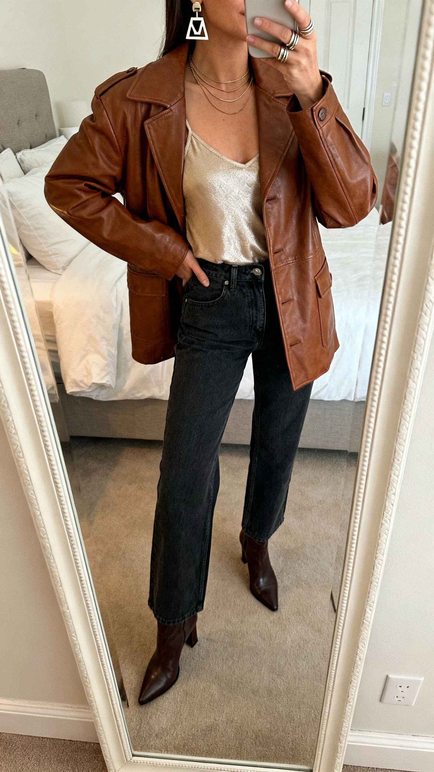iPhone mirror selfie of a woman in a classic brown leather jacket, metallic champagne cami, dark high-waisted cigarette jeans, heeled ankle boots, statement geometric earrings, thin layered necklaces and a stack of rings, face not visible, cropped at shoulders, bedroom mirror with warm indoor lighting, iPhone photo quality.