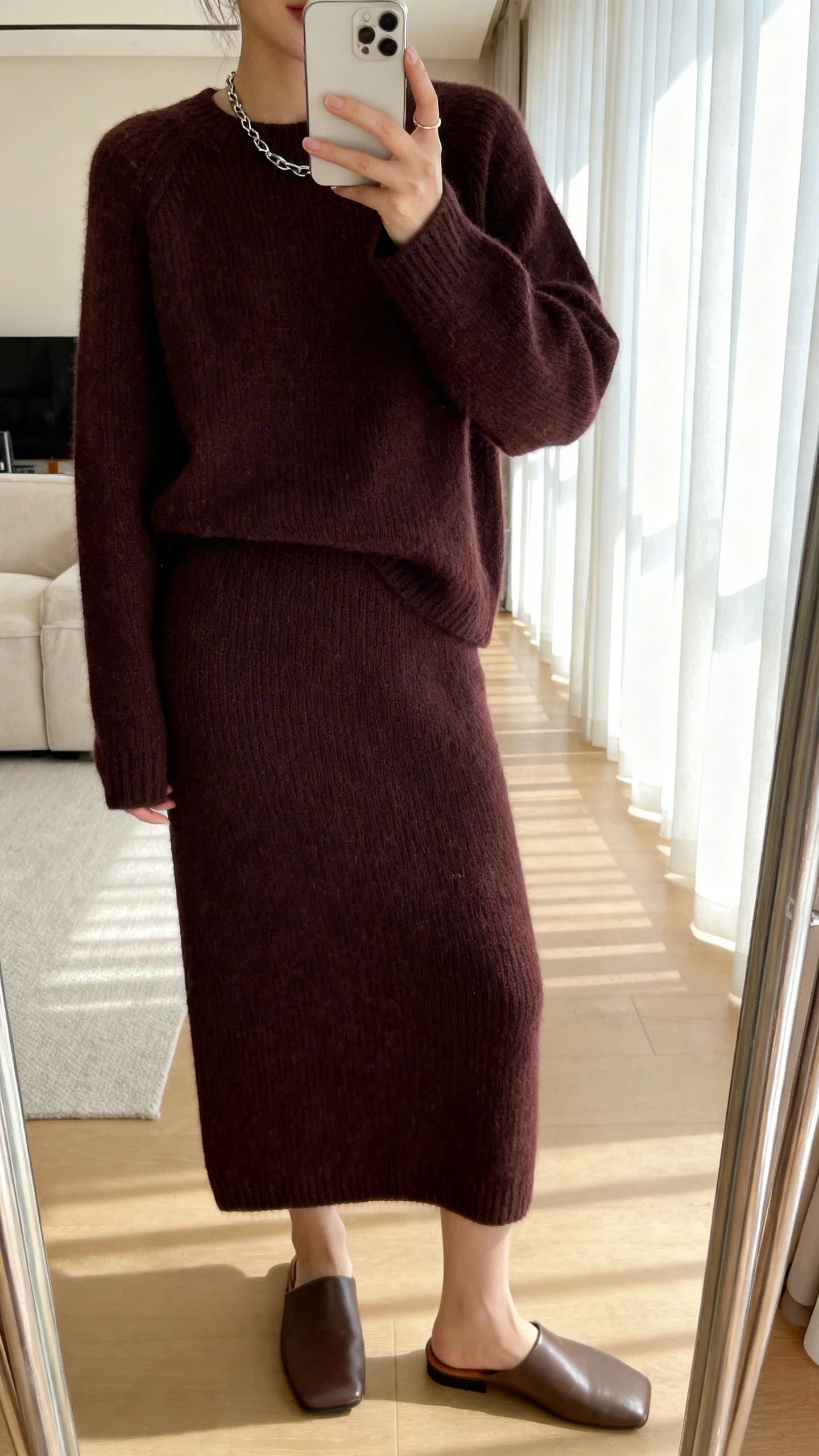 iPhone mirror selfie of a woman in a monochrome knit set (fine-knit sweater and matching knit skirt) in deep espresso, with minimalist leather mules and a subtle chain necklace, face not visible, cropped at shoulders, living room mirror with natural window light, iPhone photo quality.