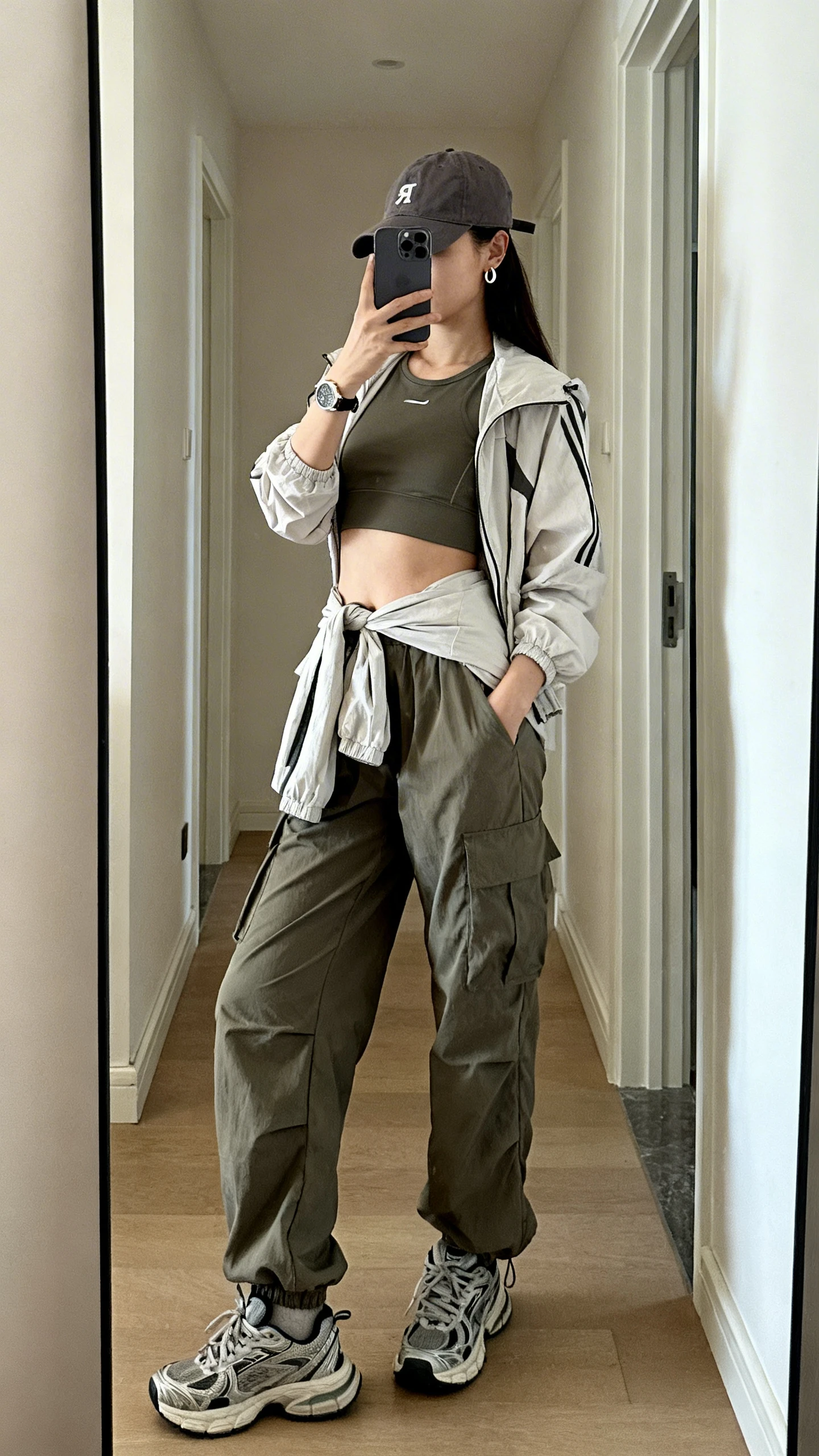 iPhone mirror selfie of a woman in a sporty-cargo fit: cropped athletic top, parachute cargo pants, chunky sneakers, baseball cap, and a lightweight windbreaker tied at the waist, with small huggie earrings and a wristwatch, face not visible, side angle, hallway full-length mirror, natural daylight, iPhone photo quality.