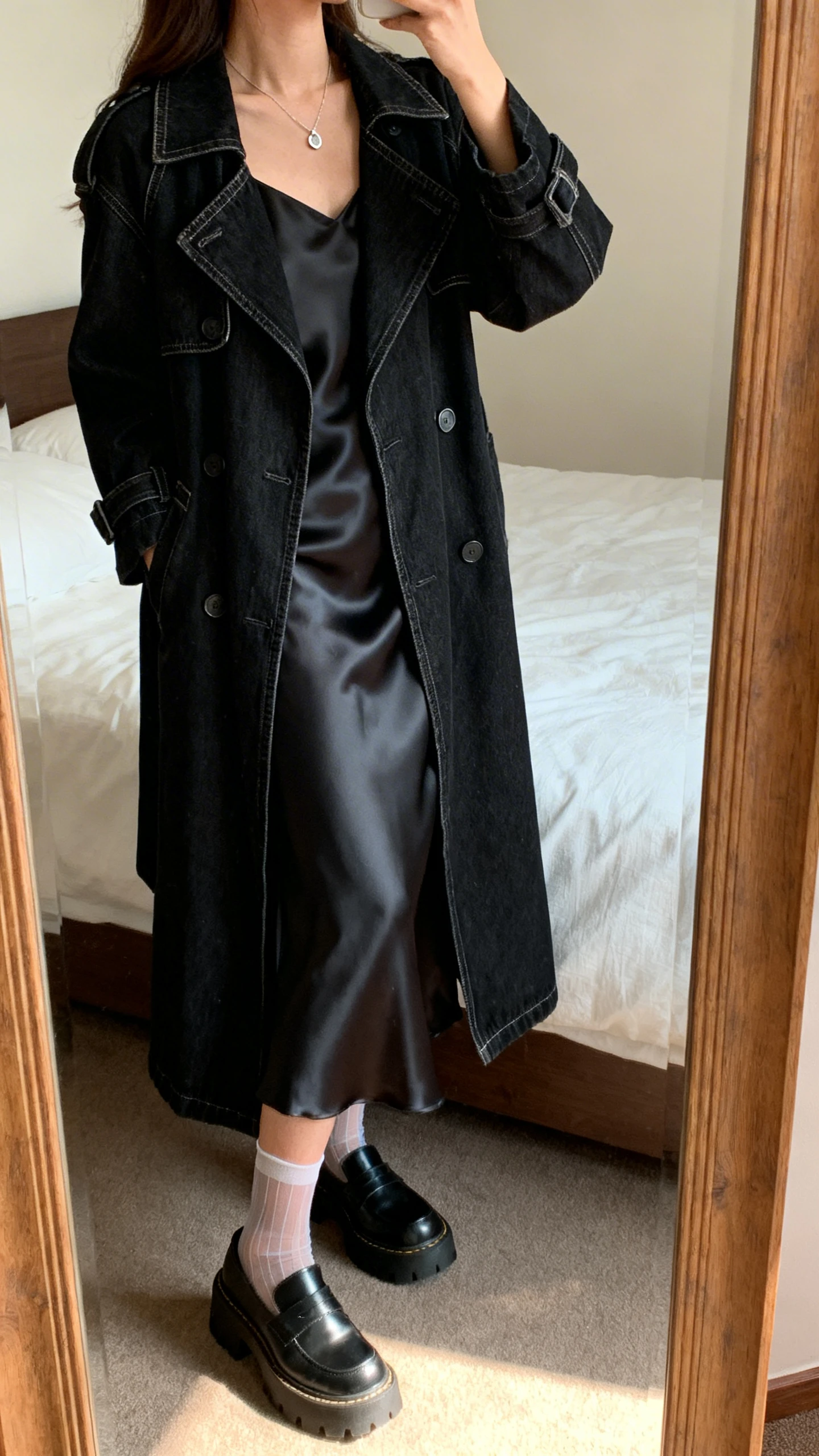 iPhone mirror selfie of a woman wearing a black denim trench layered over a satin slip dress, paired with chunky Mary Janes, sheer socks, and a delicate pendant necklace, face not visible, side angle, bedroom mirror, warm indoor lighting, iPhone photo quality.