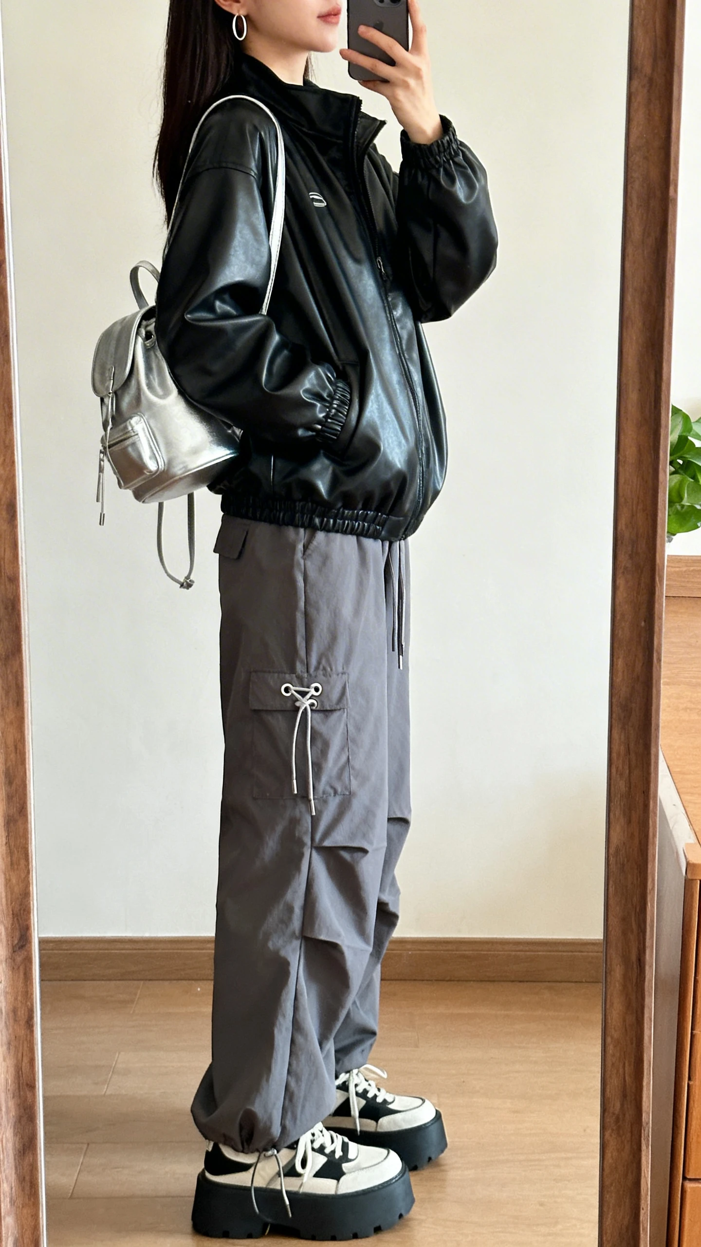 iPhone mirror selfie of a woman wearing a black leather-look windbreaker, gray parachute pants with toggle ties, platform sneakers, a silver mini backpack, and slim hoop earrings, face not visible, side angle, warm indoor lighting, iPhone photo quality.