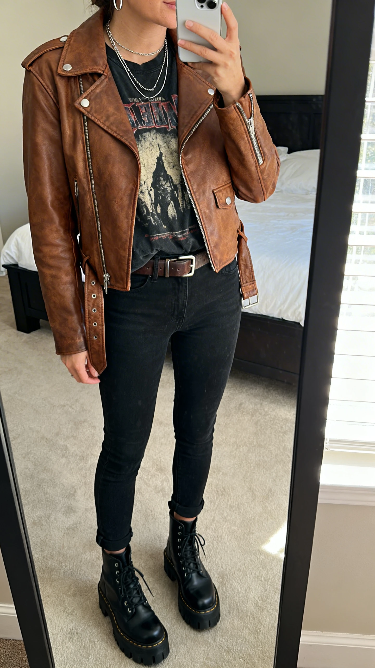 iPhone mirror selfie of a woman wearing a brown leather moto jacket, faded graphic band tee in washed black, black slim jeans slightly cropped, chunky black lace-up boots, a leather belt with a subtle metal buckle, layered silver necklaces and thin hoop earrings, face not visible, cropped at neck, bedroom mirror with natural window light, iPhone photo quality.