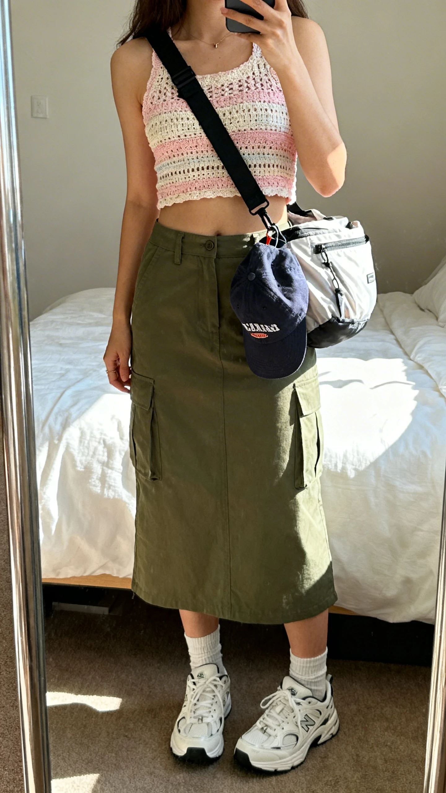 iPhone mirror selfie of a woman wearing a pastel crochet cropped tank, olive cargo midi skirt, white retro sneakers, ankle socks, a sporty nylon shoulder bag, and a baseball cap clipped to the bag, face not visible, cropped at neck, bedroom mirror with natural daylight, iPhone photo quality.