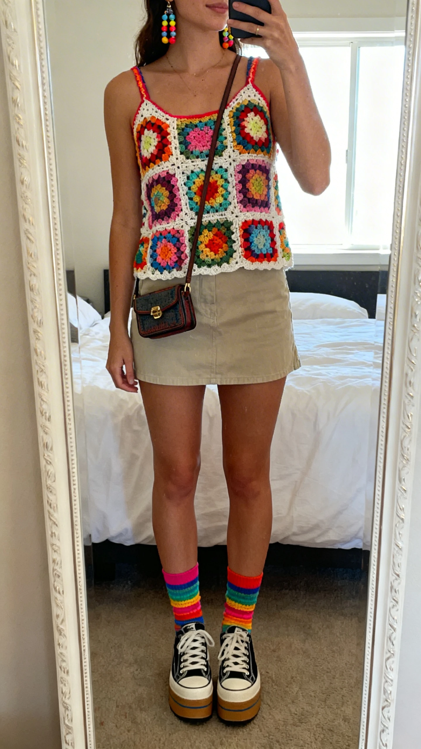 iPhone mirror selfie of a woman wearing a psychedelic granny-square crochet top with a mini skirt, colorful ankle socks with platform sneakers, beaded earrings, and a tiny shoulder bag, face not visible, cropped at shoulders, bedroom mirror, bright window light, iPhone photo quality.