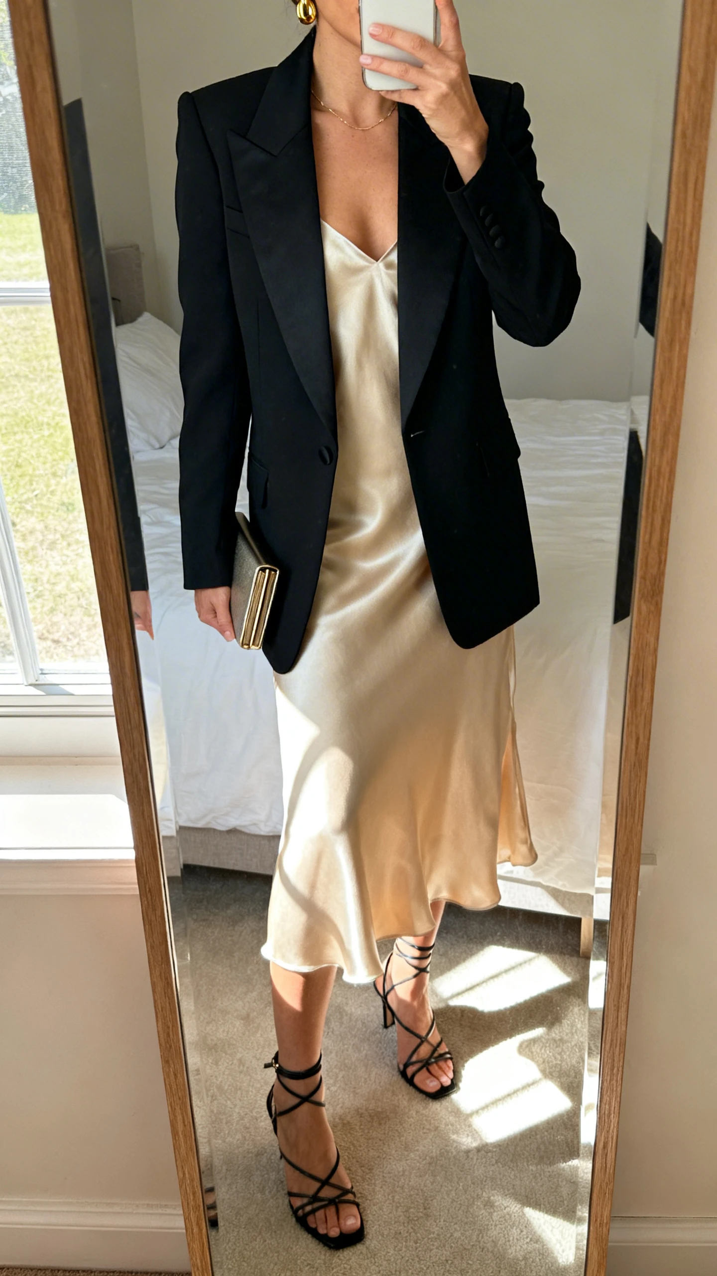 iPhone mirror selfie of a woman wearing a sharp black tuxedo blazer over a champagne silk slip dress with strappy heels, delicate gold earrings, and a slim clutch, face not visible, cropped at neck, bedroom mirror with morning window light, iPhone photo quality.
