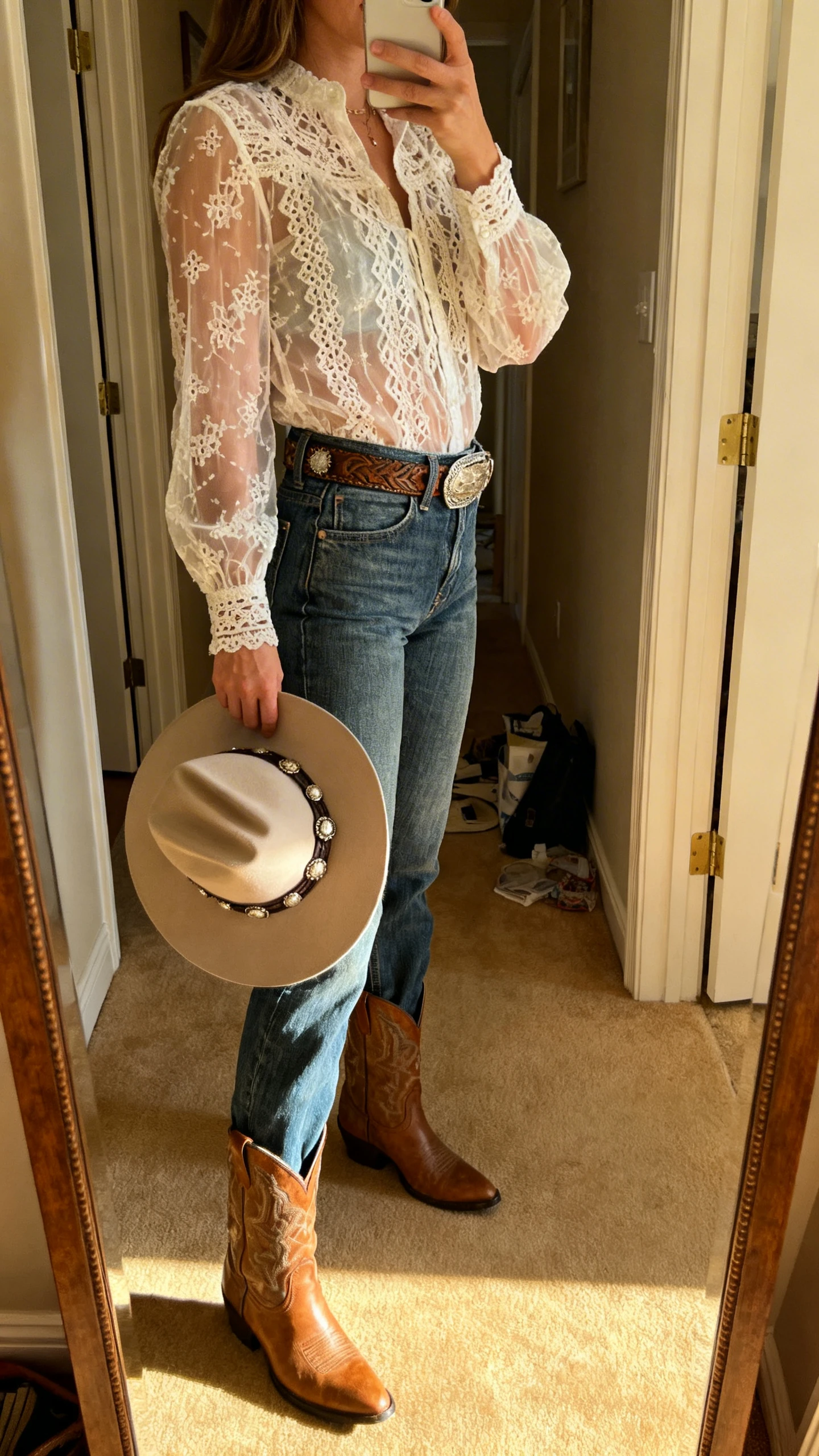 iPhone mirror selfie of a woman wearing a sheer lace-crochet blouse tucked into high-rise denim with a Western belt, ankle cowboy boots, and a concho-trim hat in hand, face not visible, side angle, entryway full-length mirror, warm indoor lighting, iPhone photo quality.